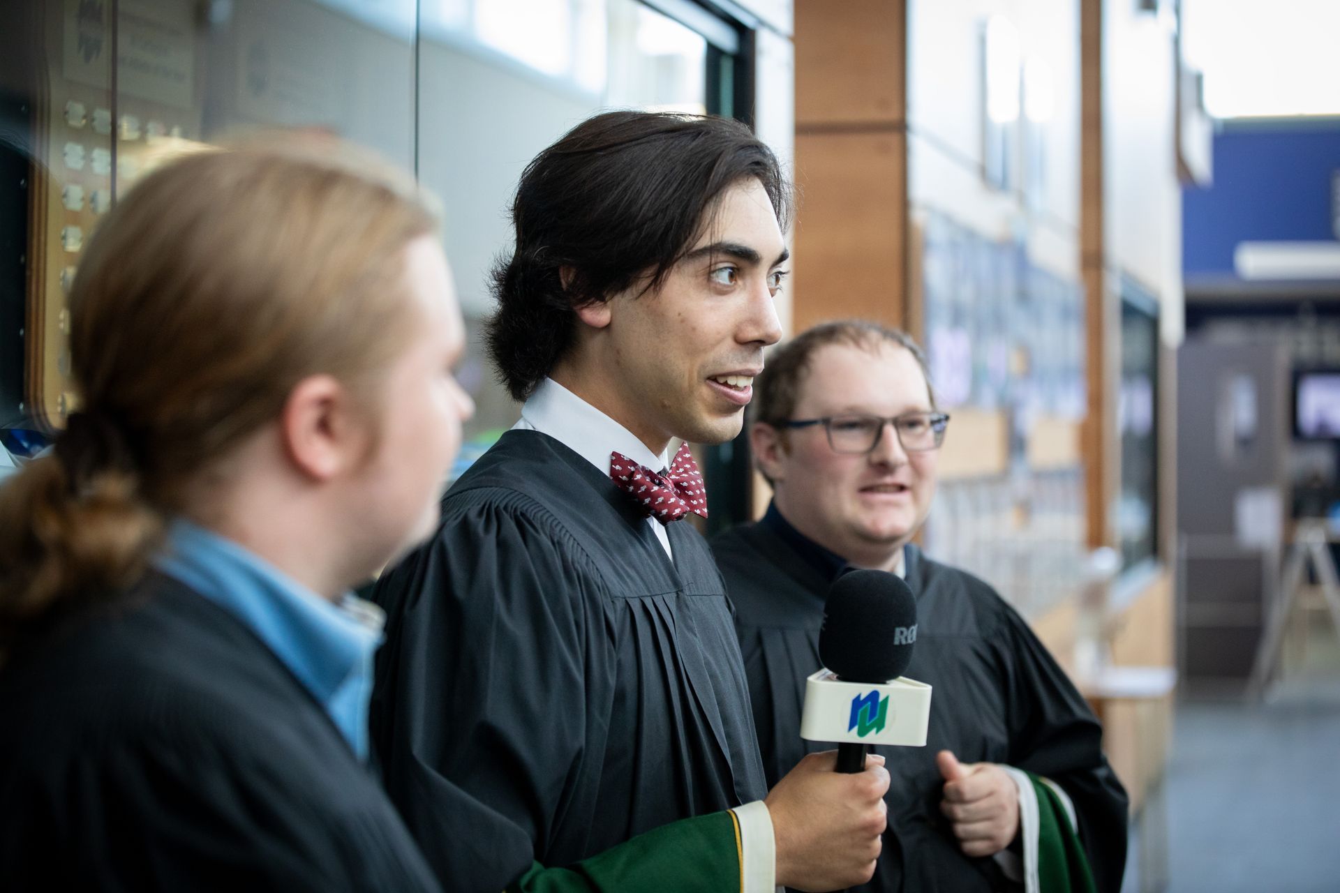 Man in graduation gown speaking into a microphone, with others nearby.