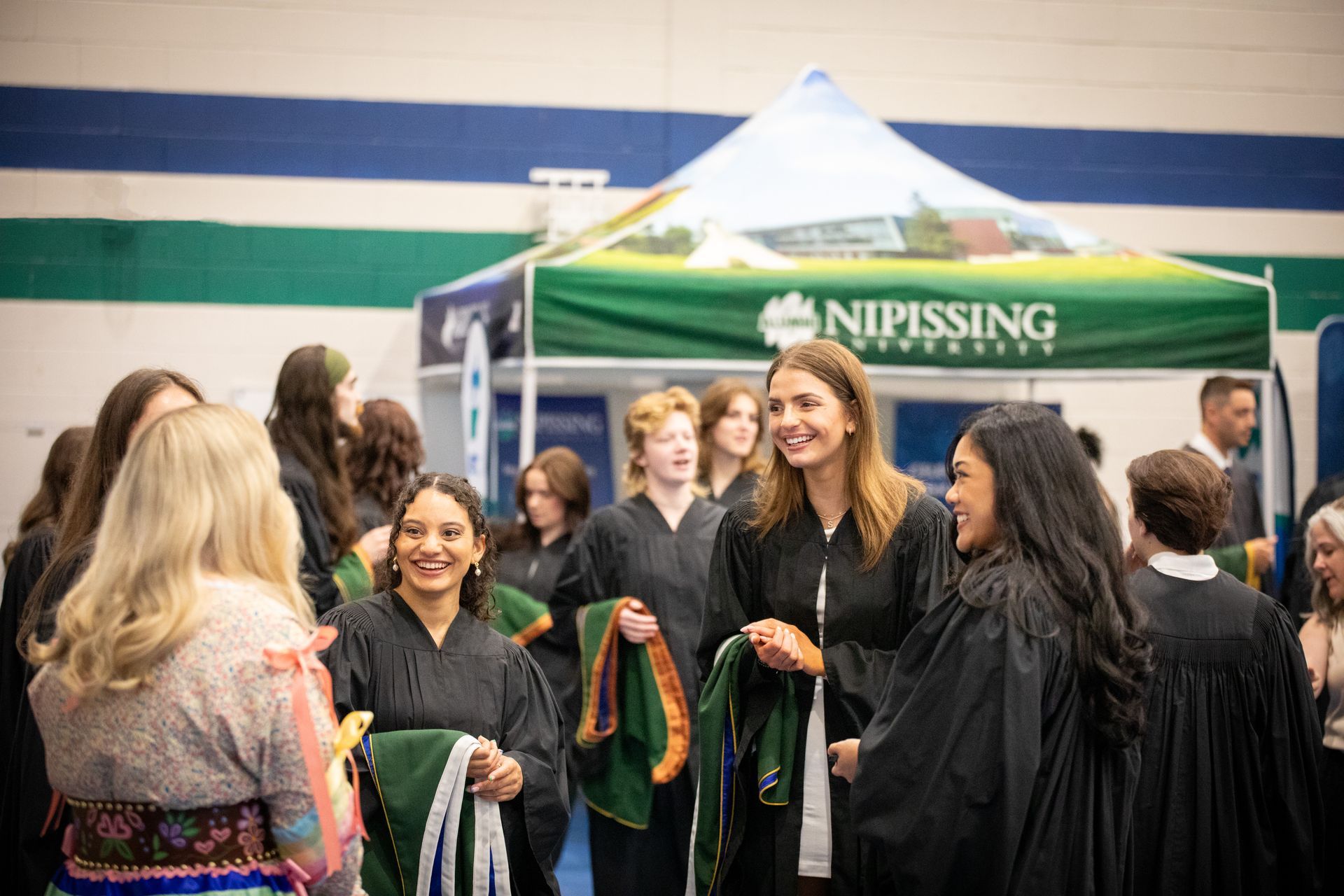 Graduates in gowns gather beside a green Nipissing tent at a ceremony booth