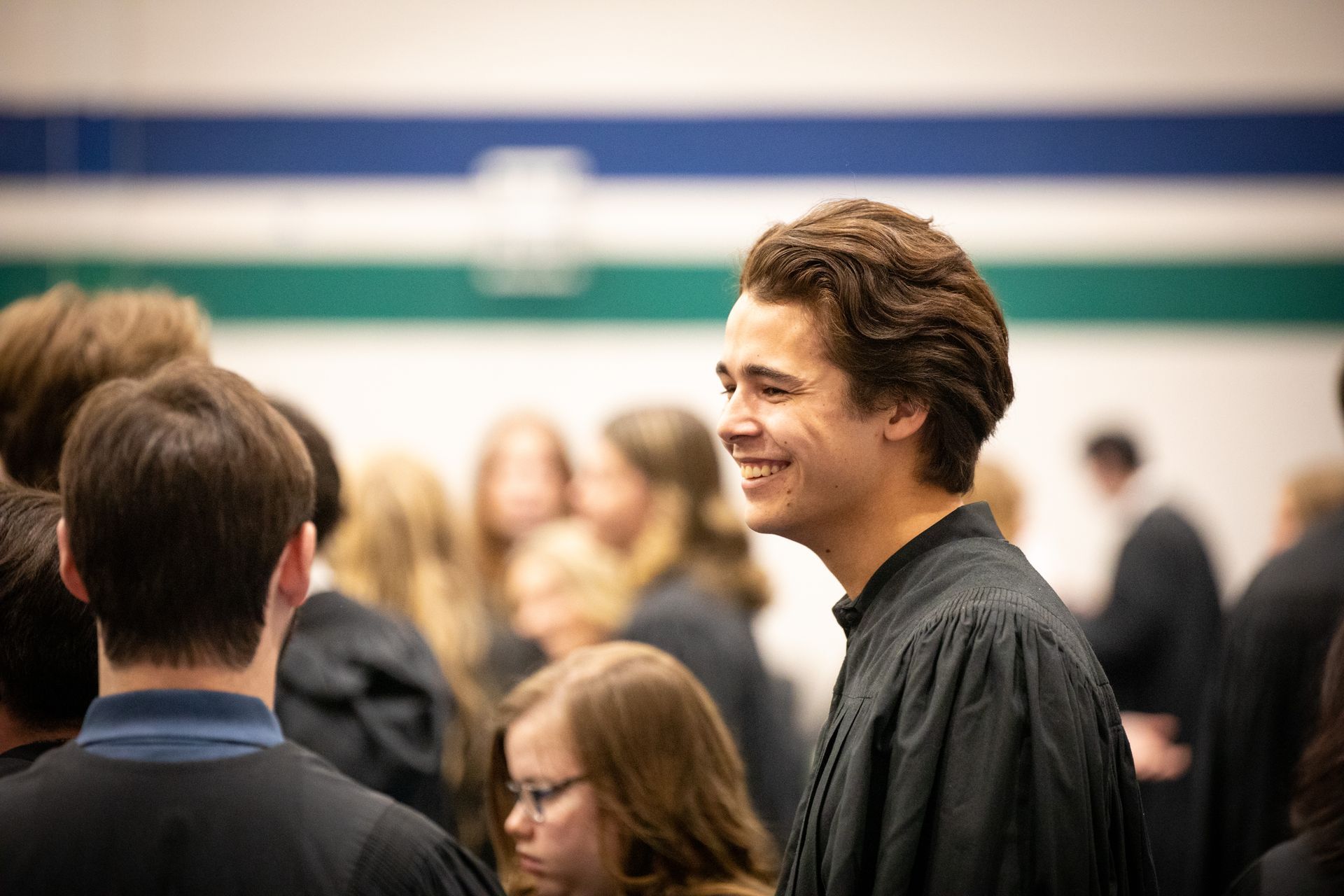 Smiling student in black gown chatting in a blurred indoor crowd.