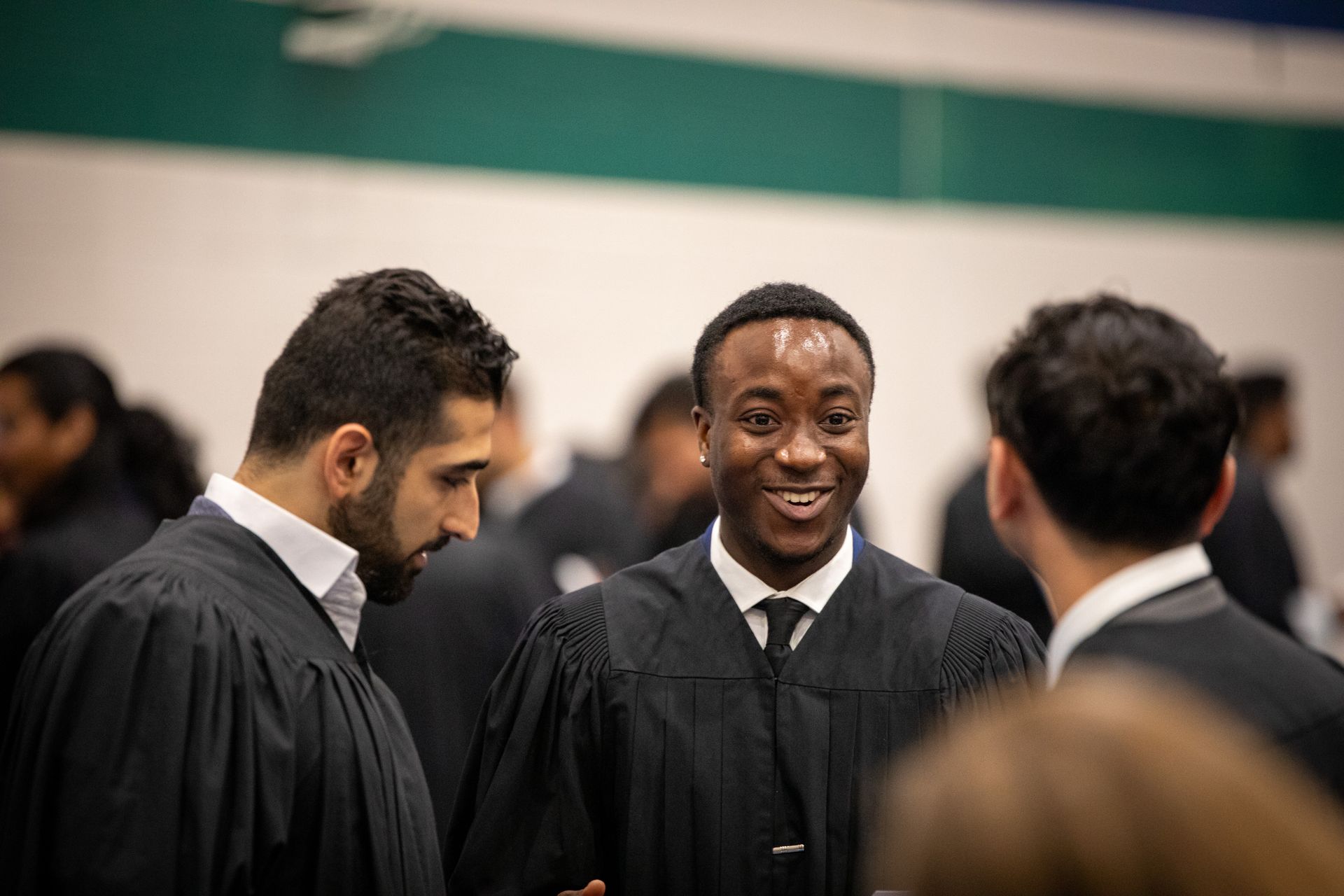 Three graduates in black gowns talking indoors, one smiling at center