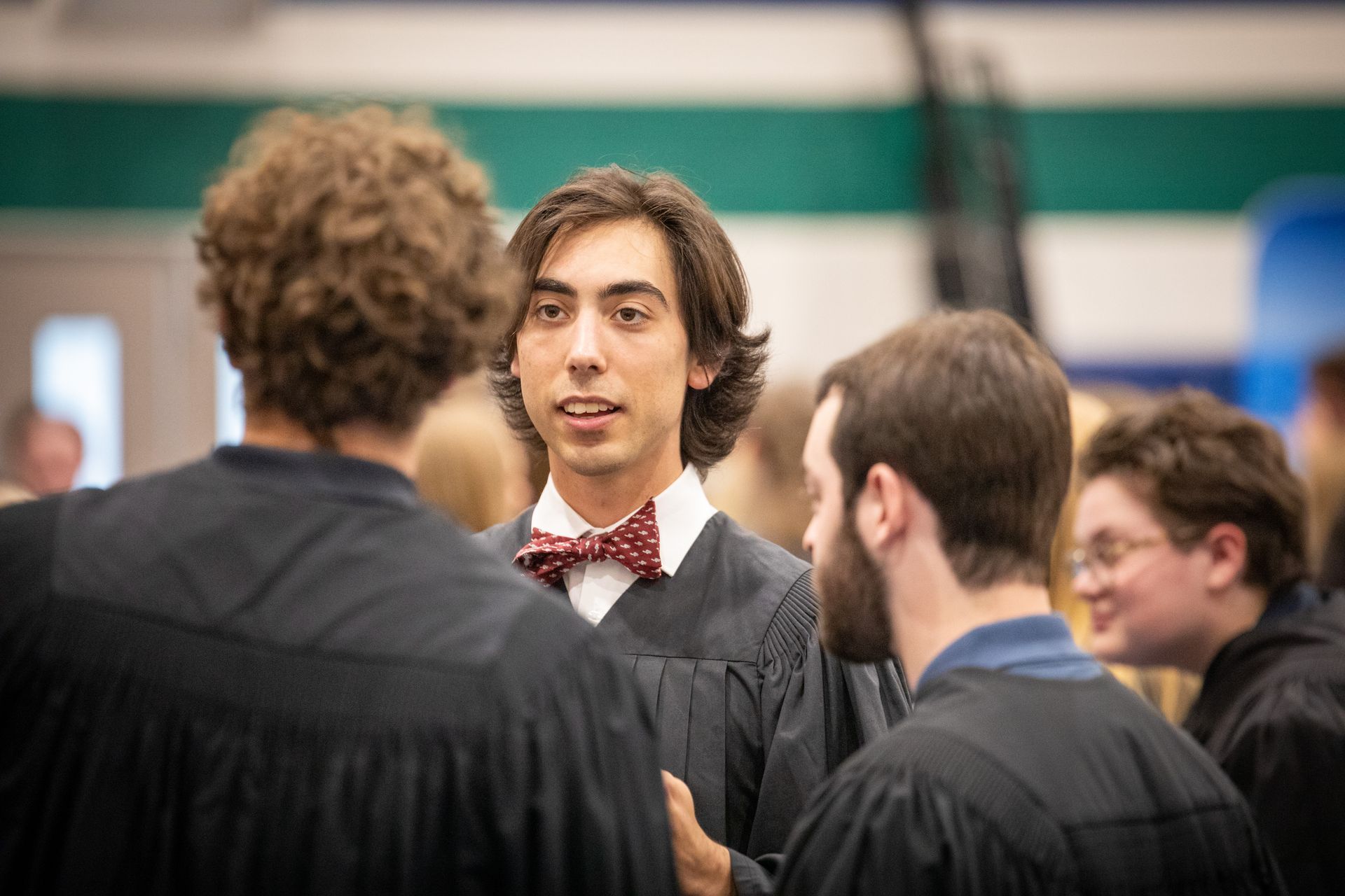 Graduate in black gown and red bow tie speaking with other graduates at a ceremony