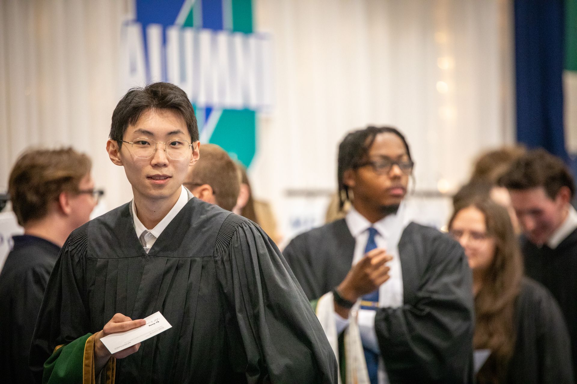 Graduate in gown holding a card, standing among people at a formal ceremony.