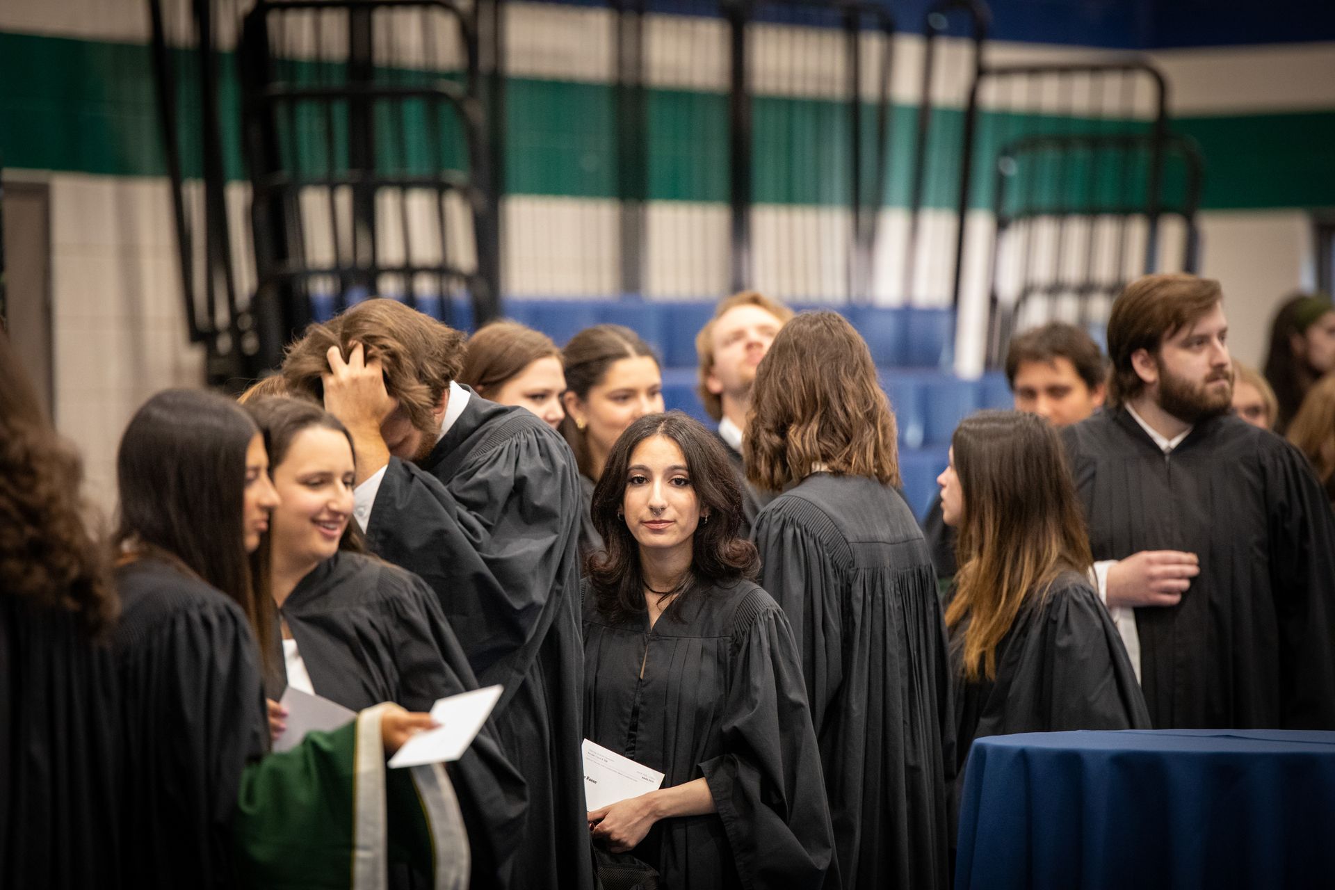 People in black graduation gowns mingling indoors near draped curtains and a table