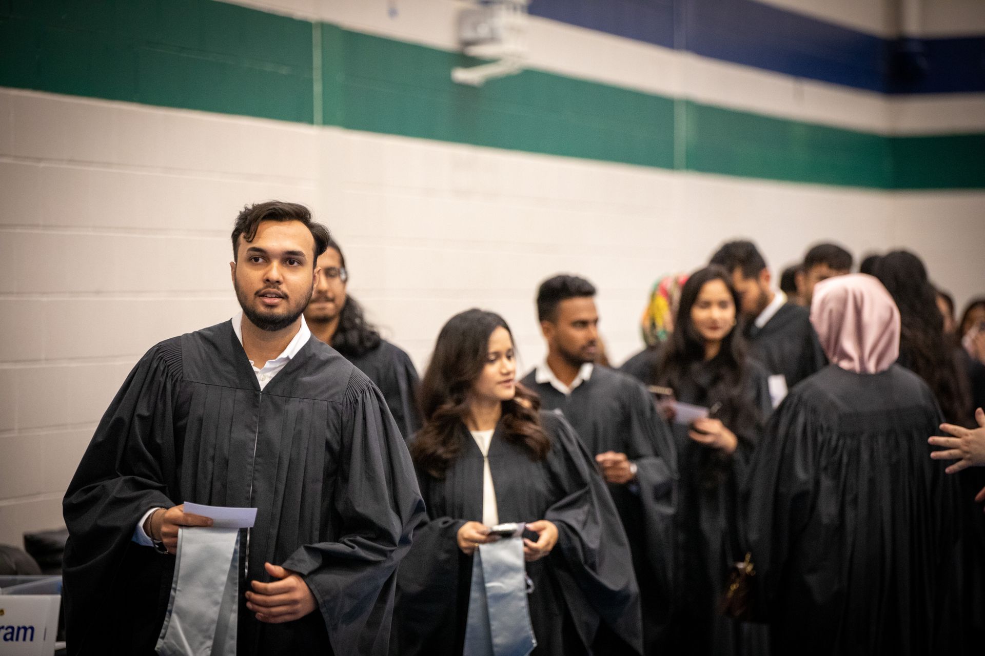 Graduates in black gowns stand in line indoors before a ceremony