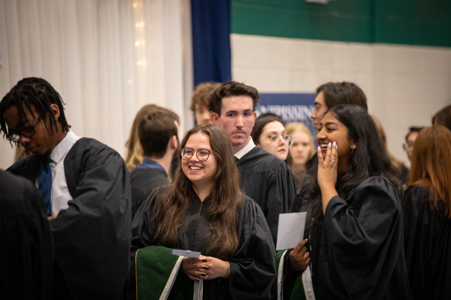 Graduates in black gowns stand together indoors, smiling and holding papers at a ceremony