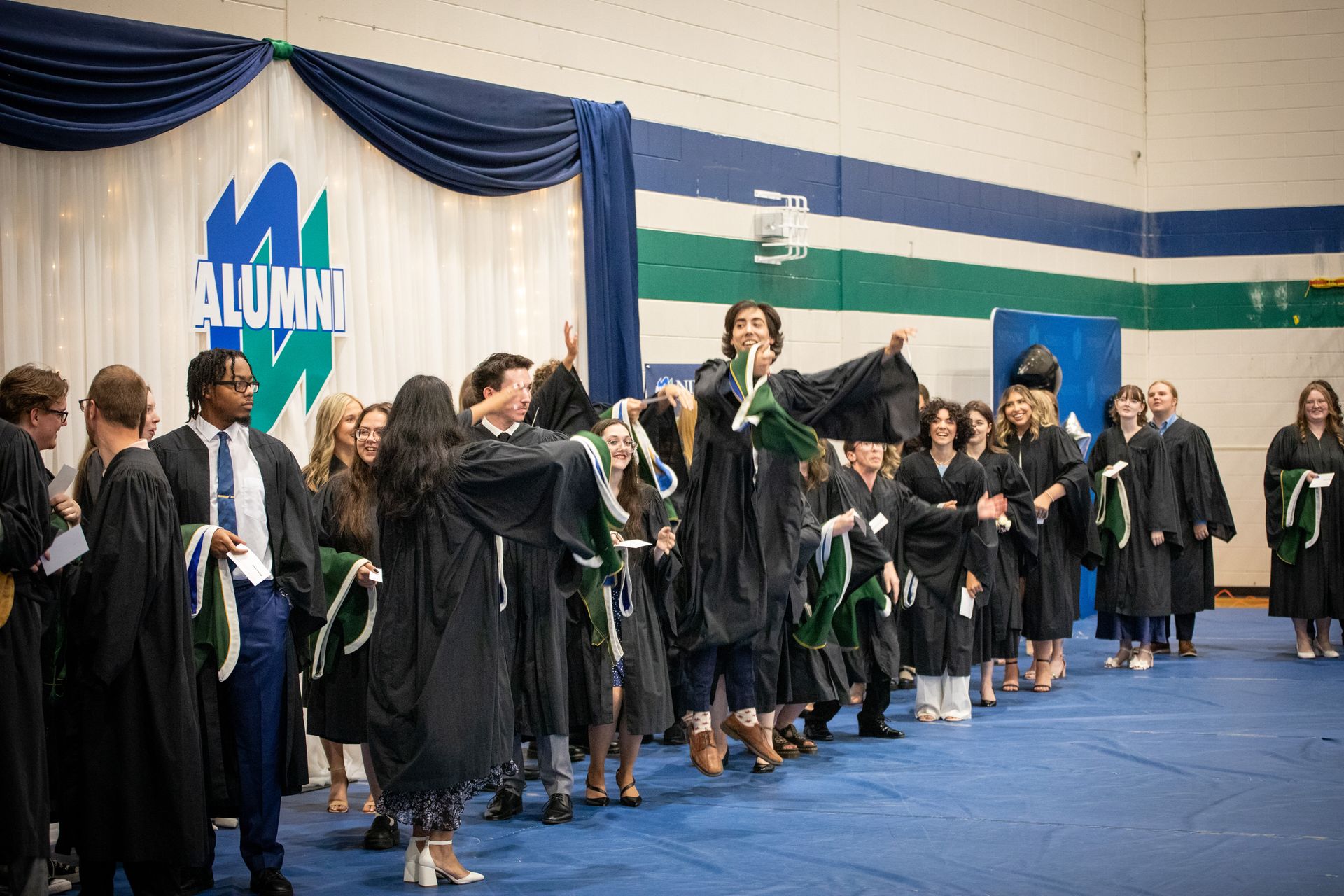Graduates in gowns line up on a blue gym floor during a commencement ceremony, some jump