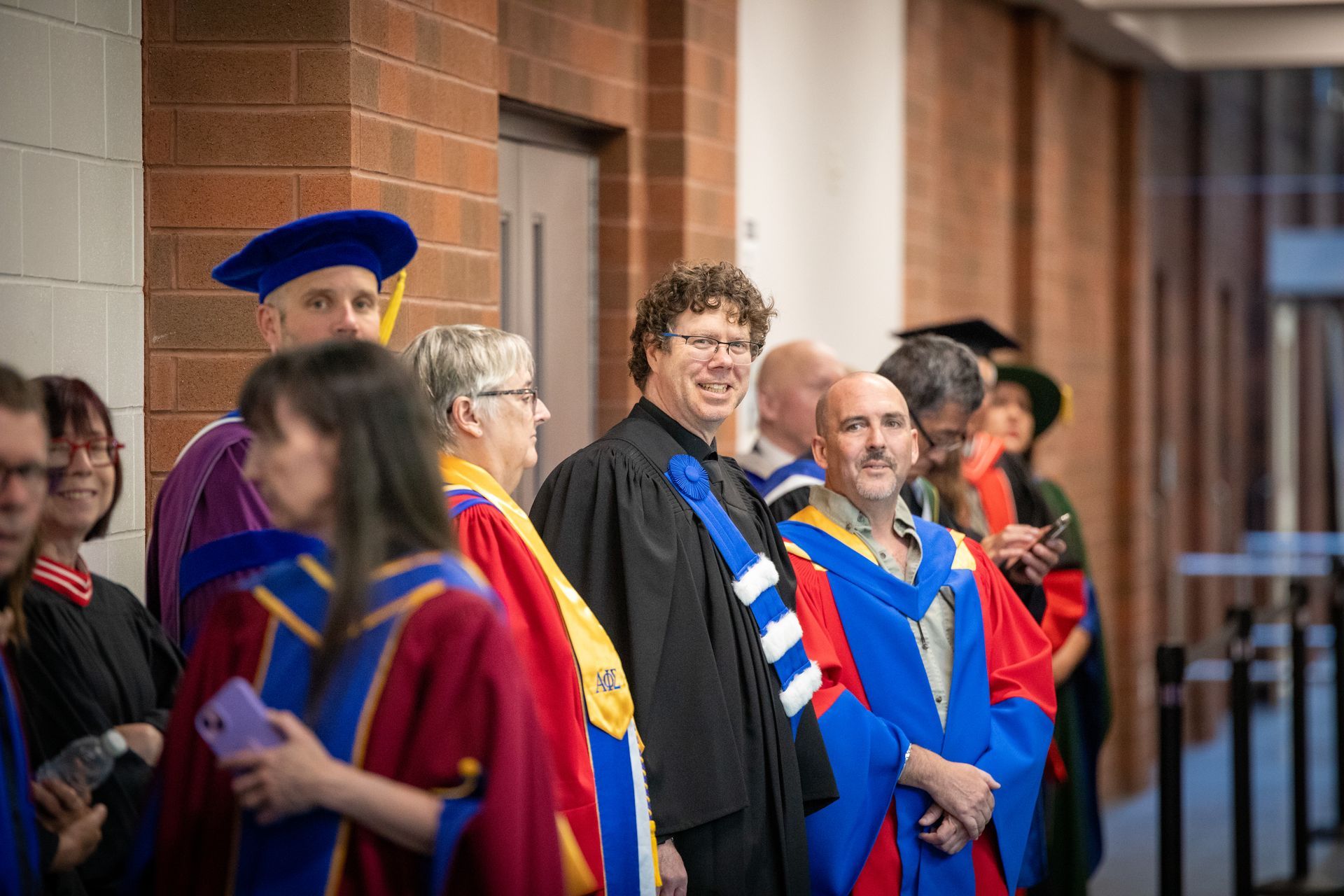 Faculty in blue and red academic gowns standing together in a brick-lined corridor