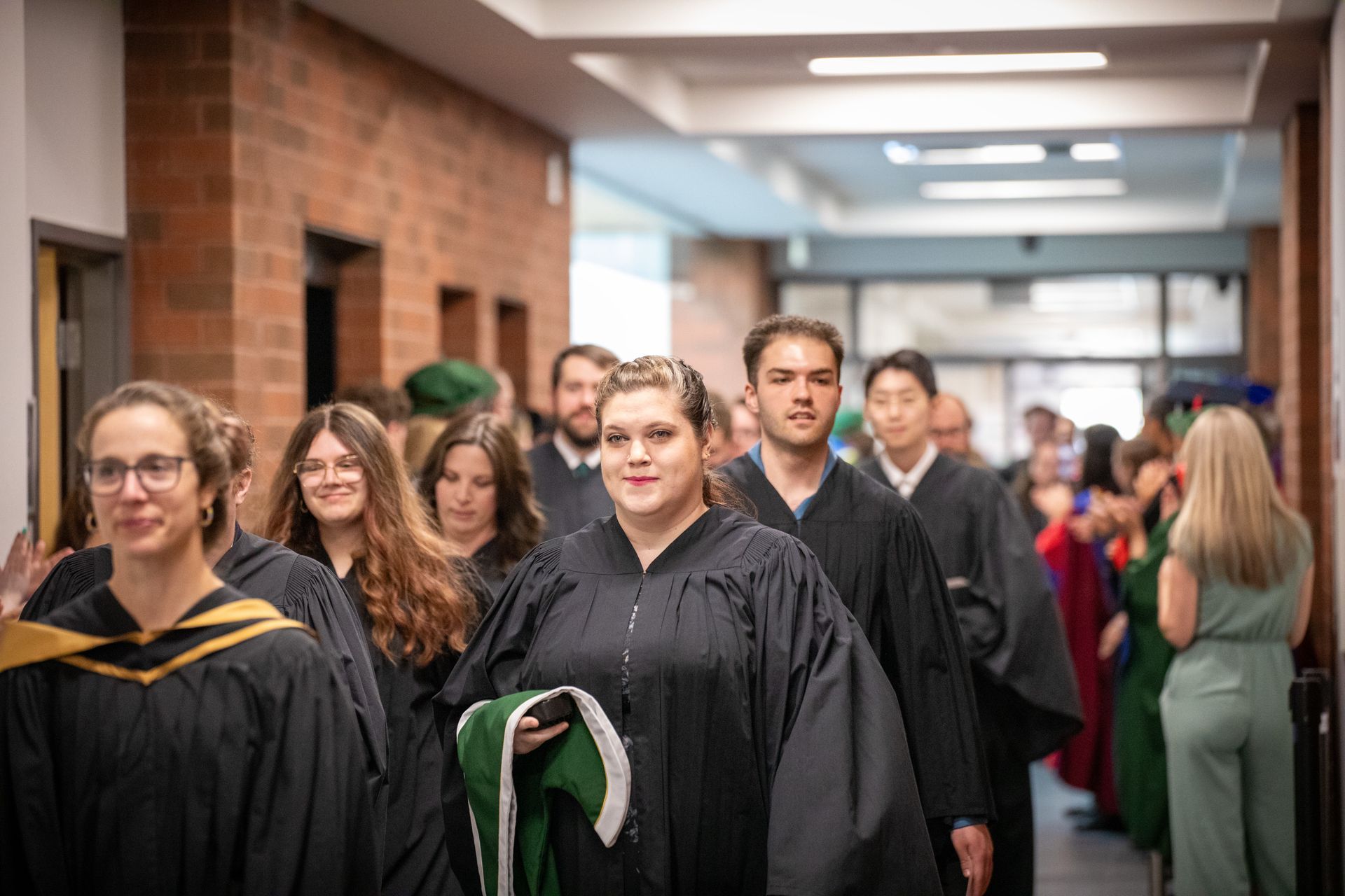 Graduates in black gowns walk through a school hallway during a ceremony processional