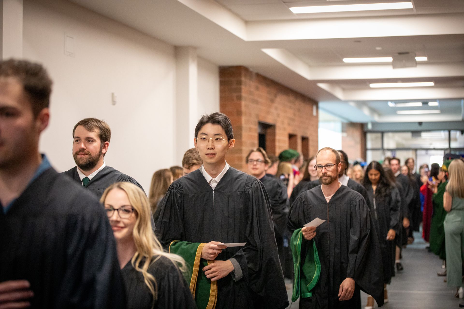 Graduates in black gowns walking down a hallway, some carrying green sashes.