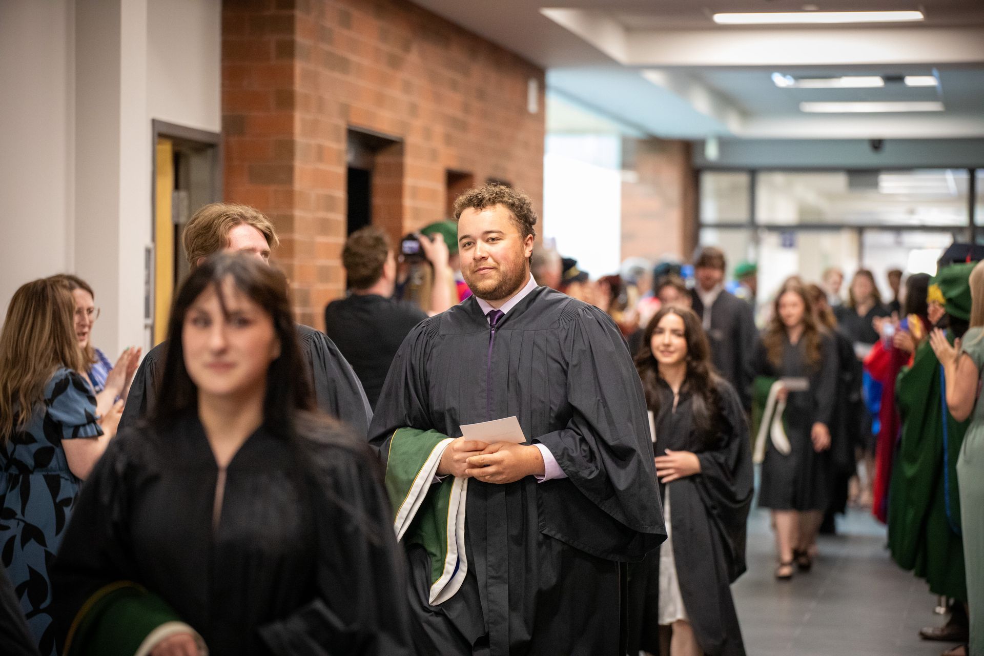 Graduates in black gowns line a hallway during a commencement procession