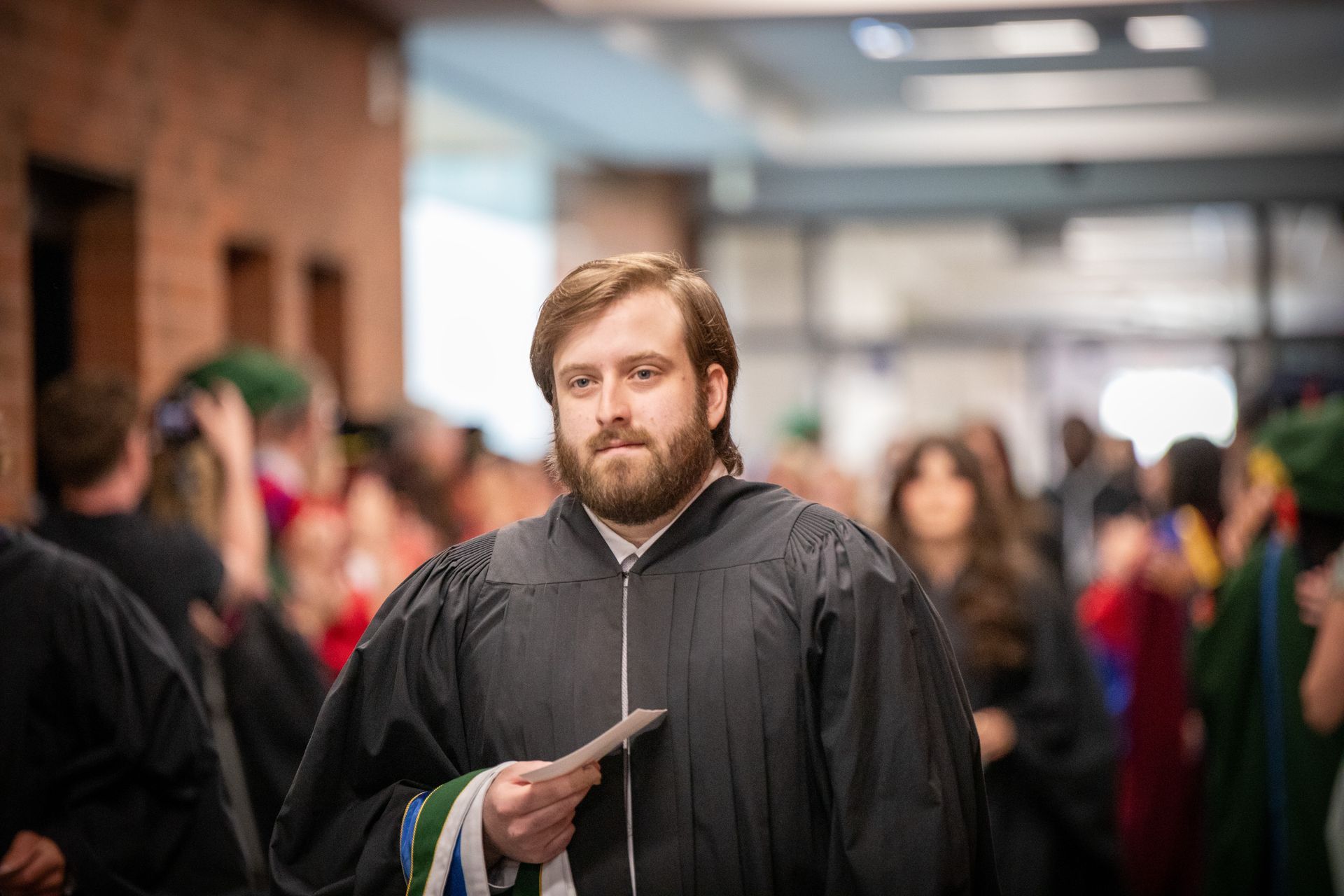 Graduate in black gown holding papersin a hallway, with blurred crowd in the background