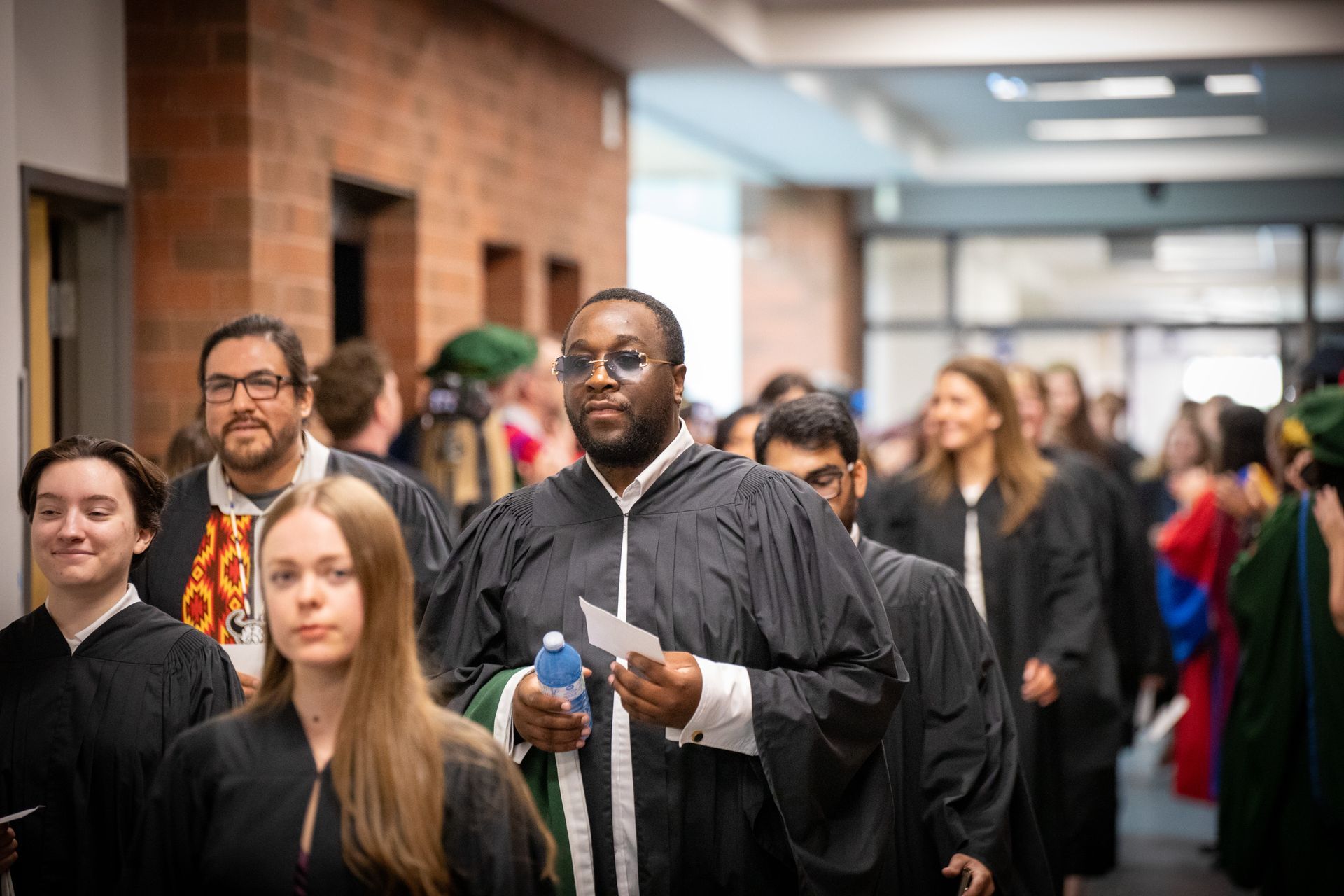 People walking through a hallway, including one person in a black gown holding papers.