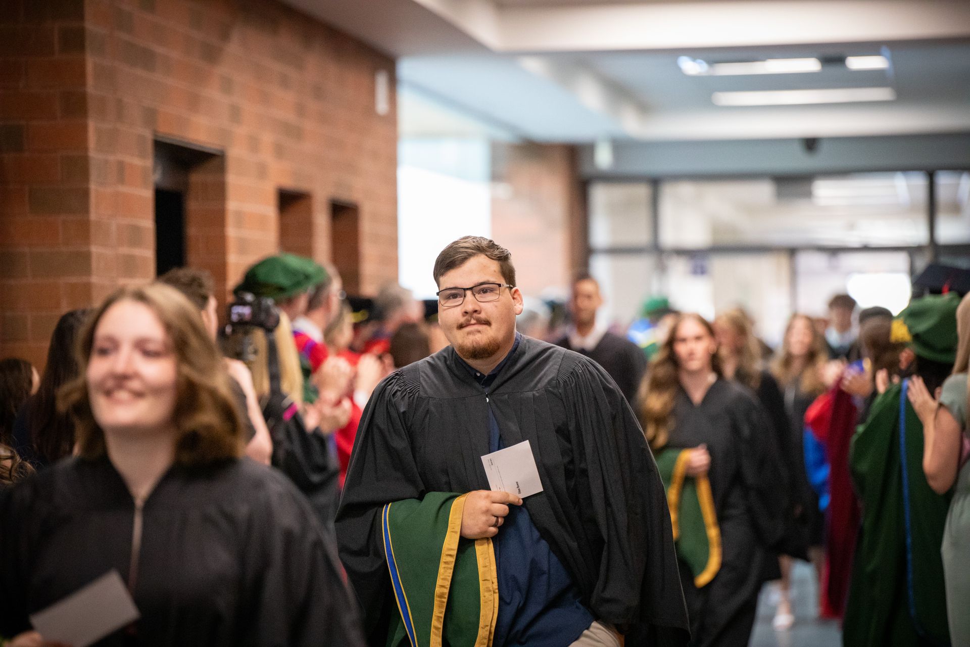 Graduates in black gowns walking through a hallway at a ceremony, holding papers