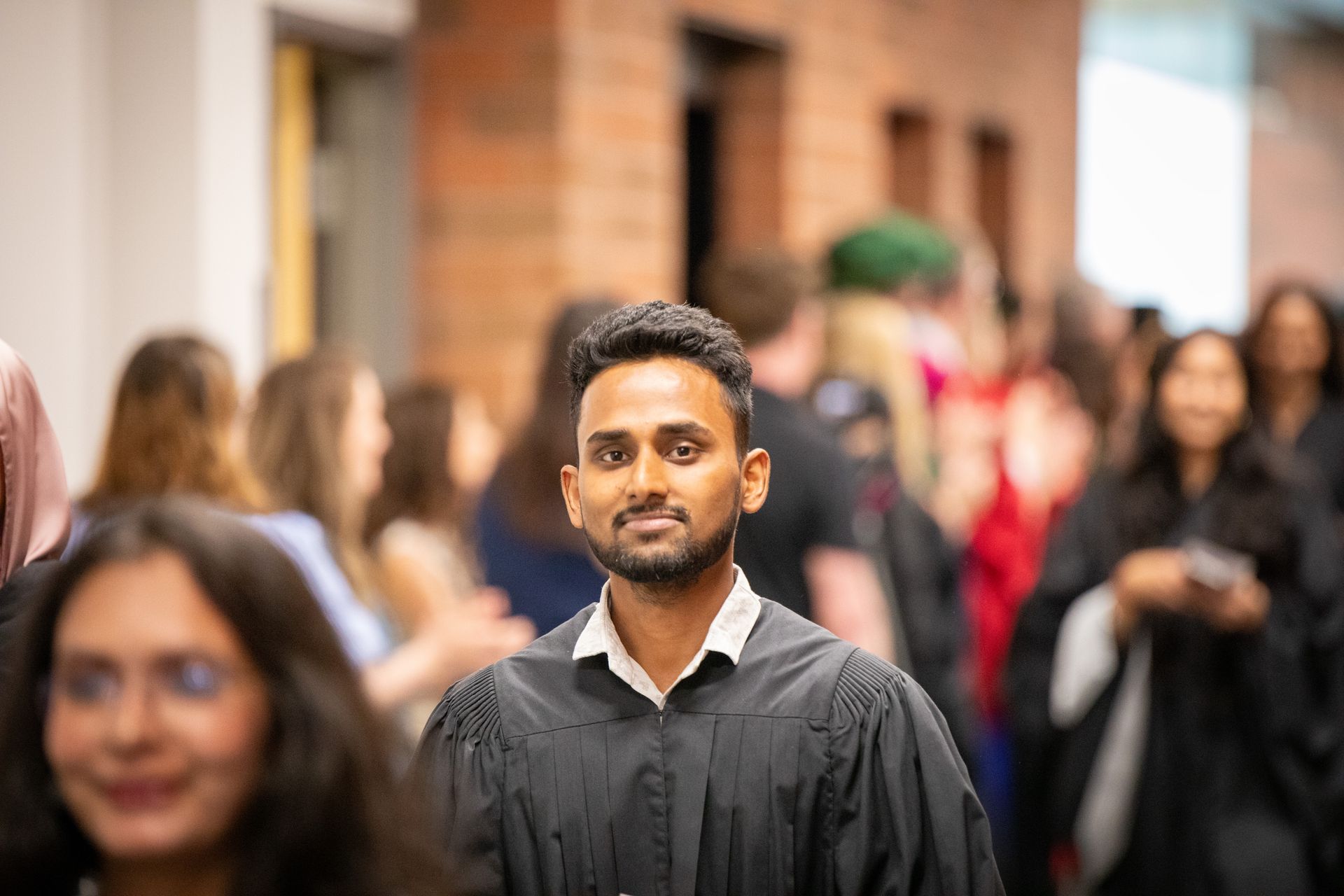 Graduate in black gown standing in a blurred crowd at a ceremony