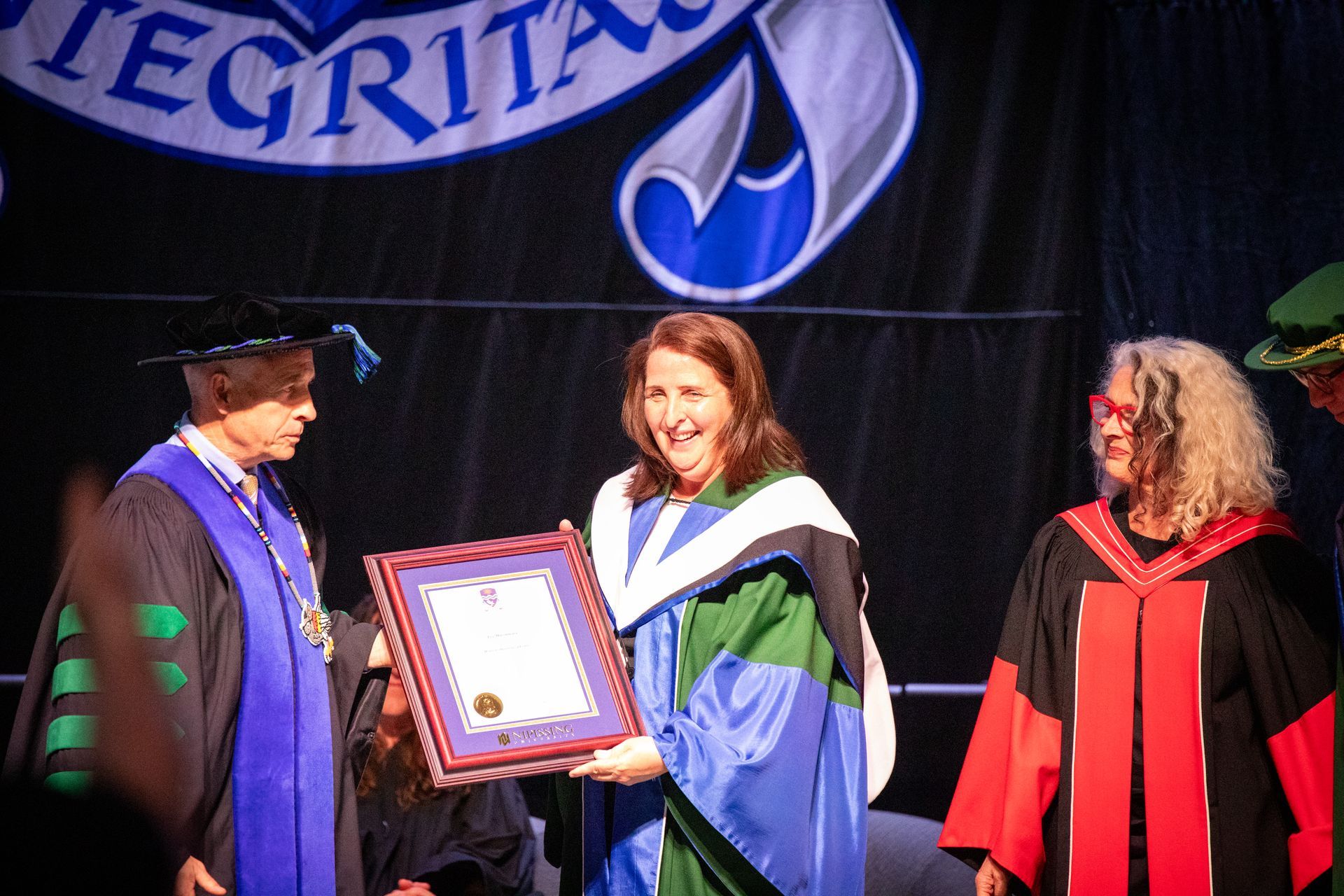 Graduation ceremony with a woman receiving a framed award on stage from two robed officials