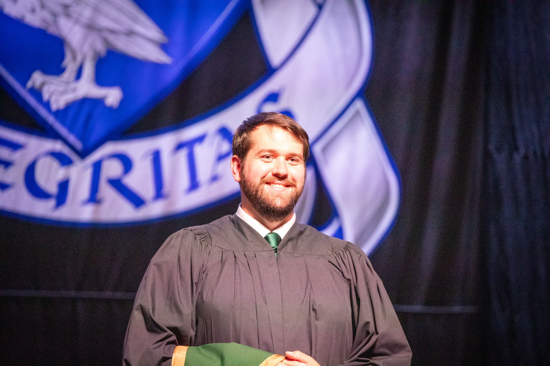 Graduate in black gown stands on stage before a blue banner, holding a diploma folder.