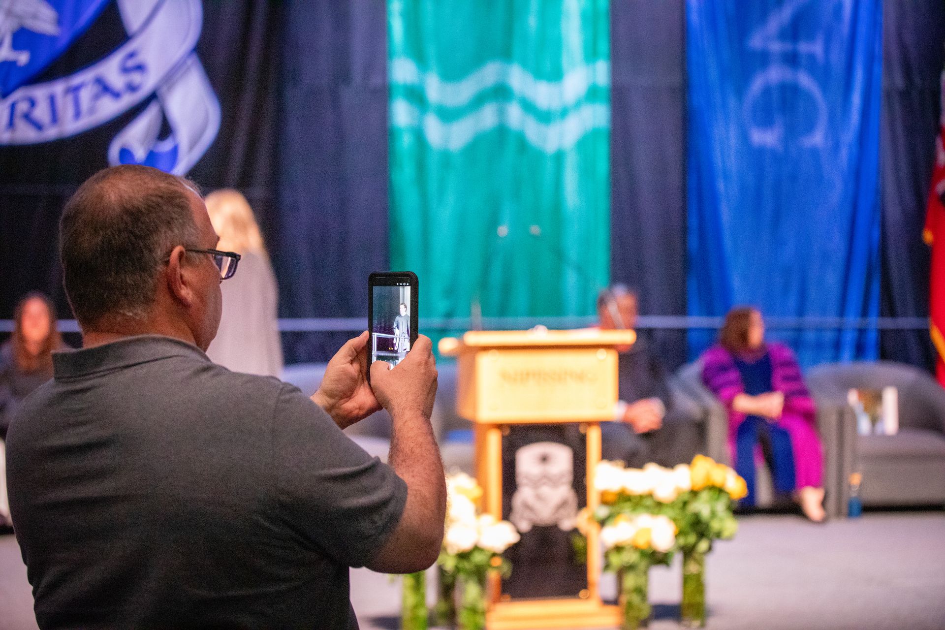 Man filming a stage ceremony with a smartphone, speaker seated beside a podium with flowers and banners behind.