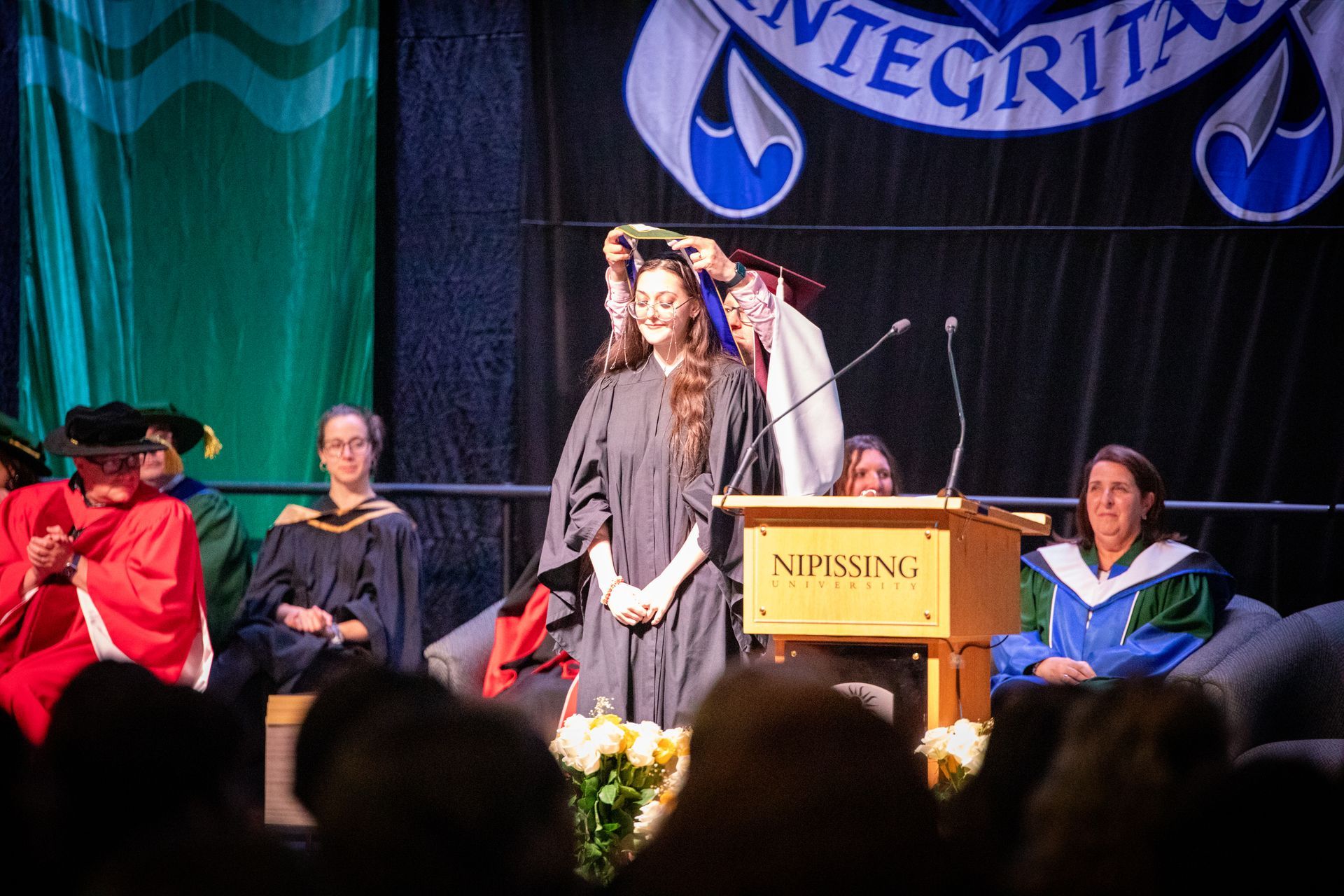 Person at a podium during a graduation ceremony on stage with seated faculty behind them