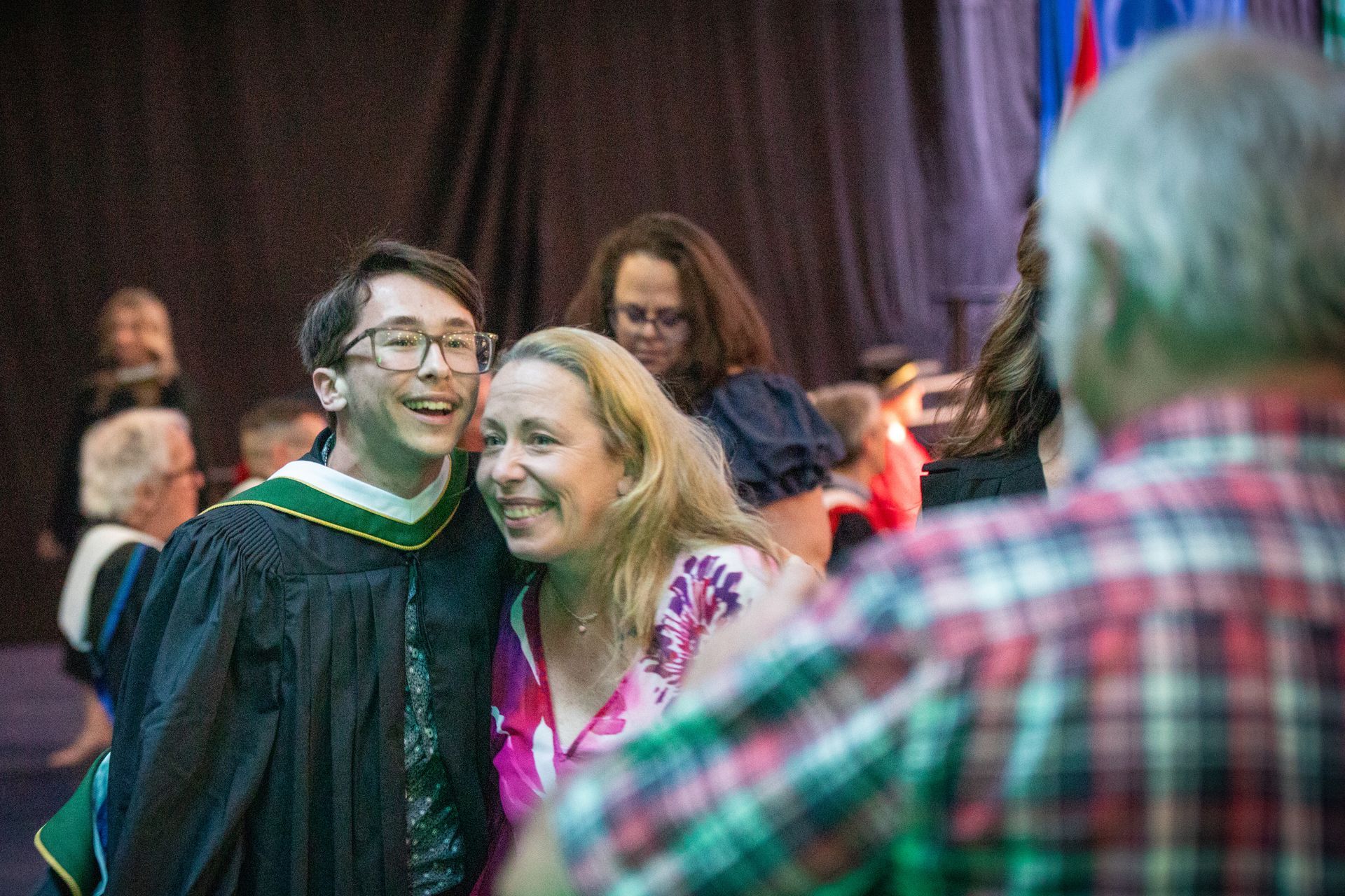 Smiling graduate in black gown and green hood posing with a woman at a ceremony, with a blurred audience behind them