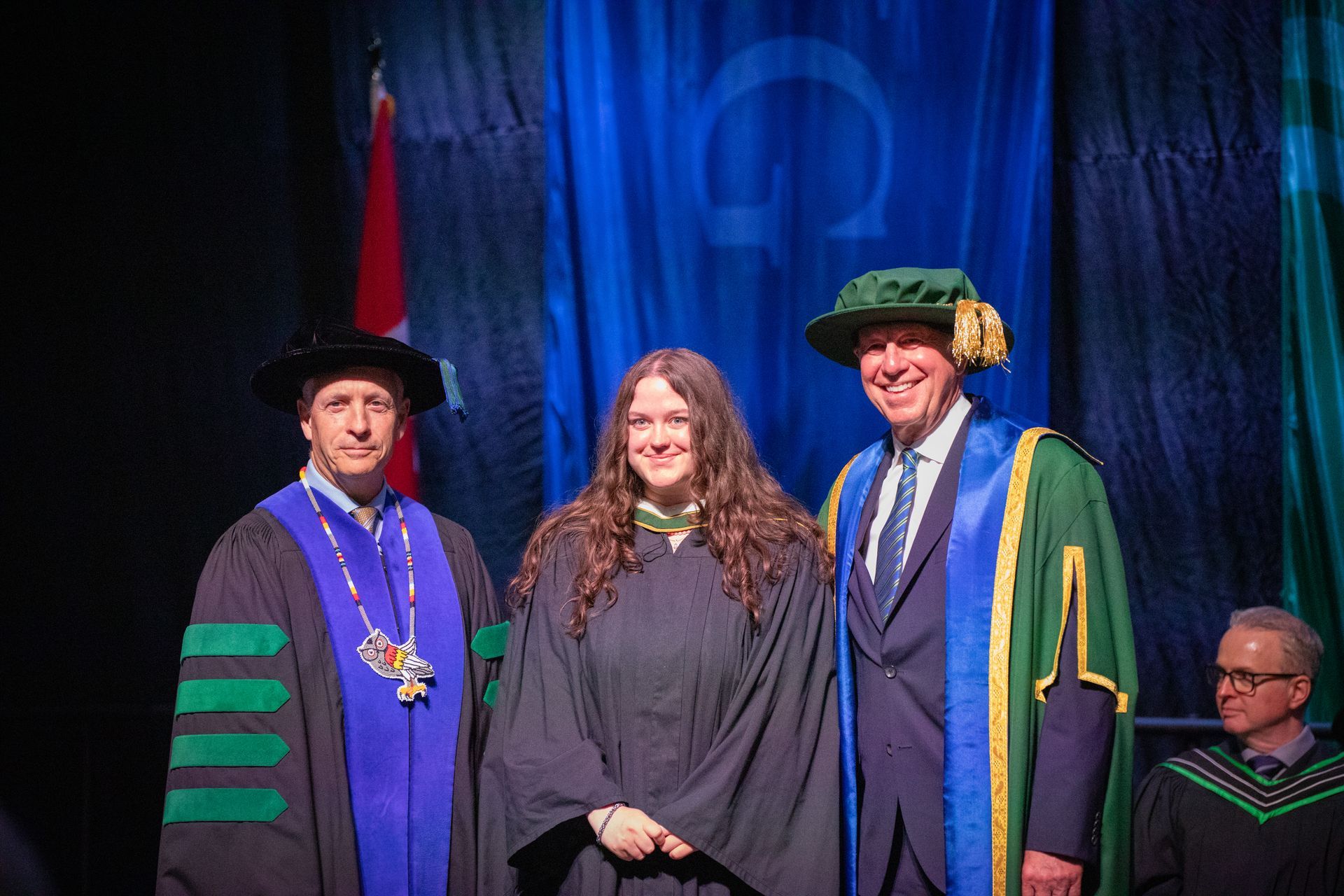 Three people in academic regalia on a stage, with blue curtains and a podium backdrop