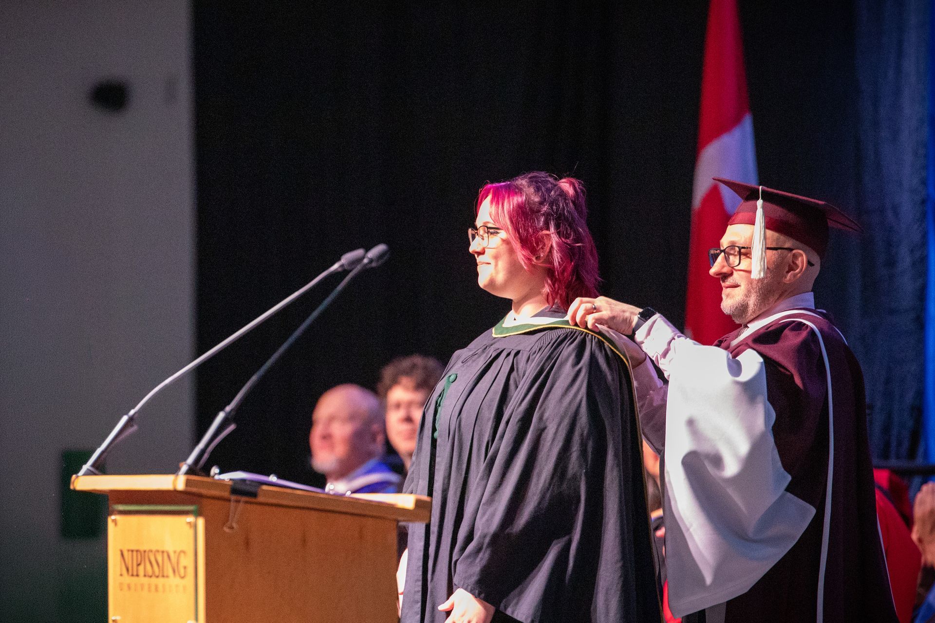 Graduation speaker placing a hood on a smiling graduate at a podium with a microphone