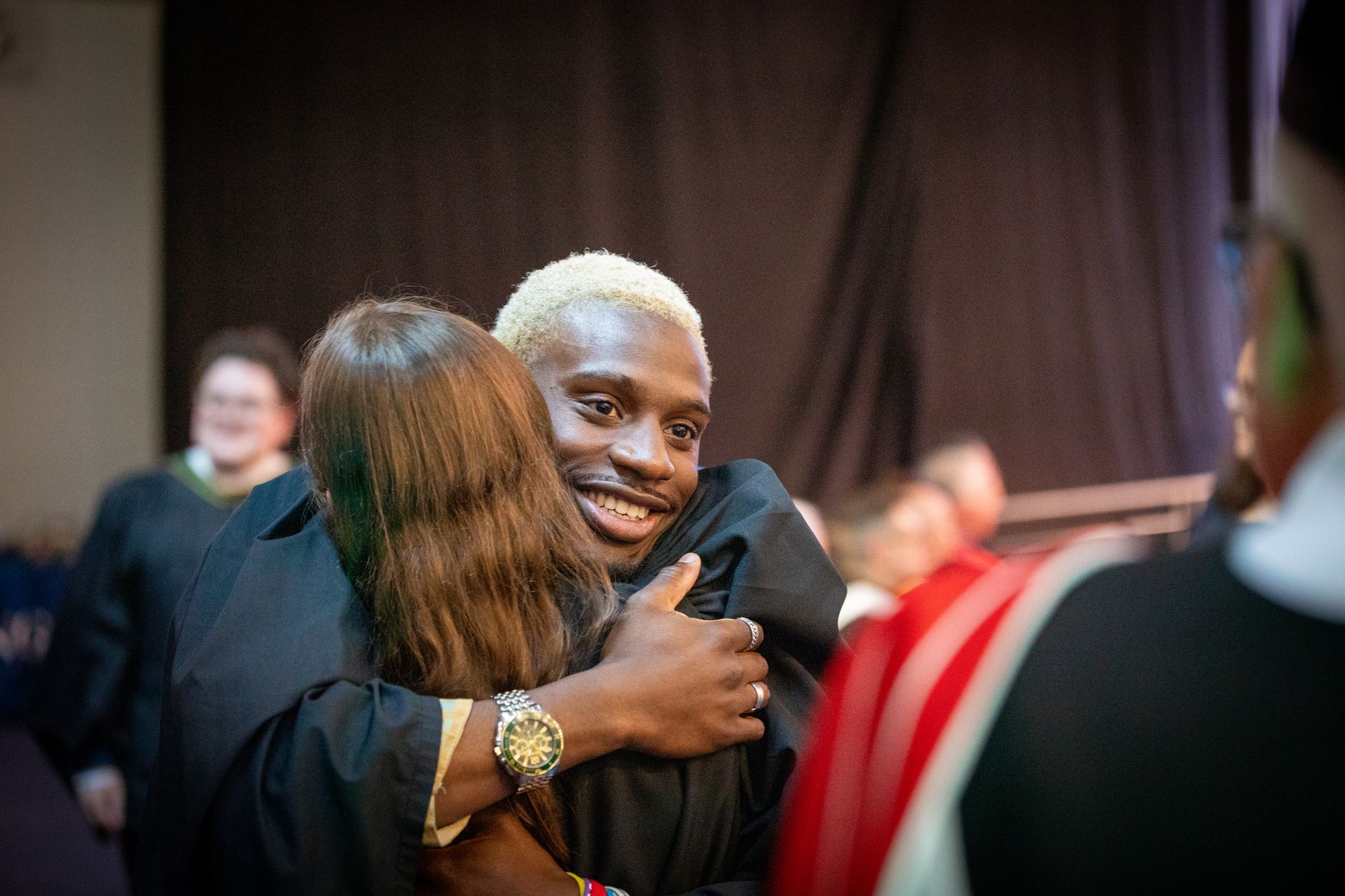 Two people hugging and smiling at an indoor event, with blurred attendees in the background.