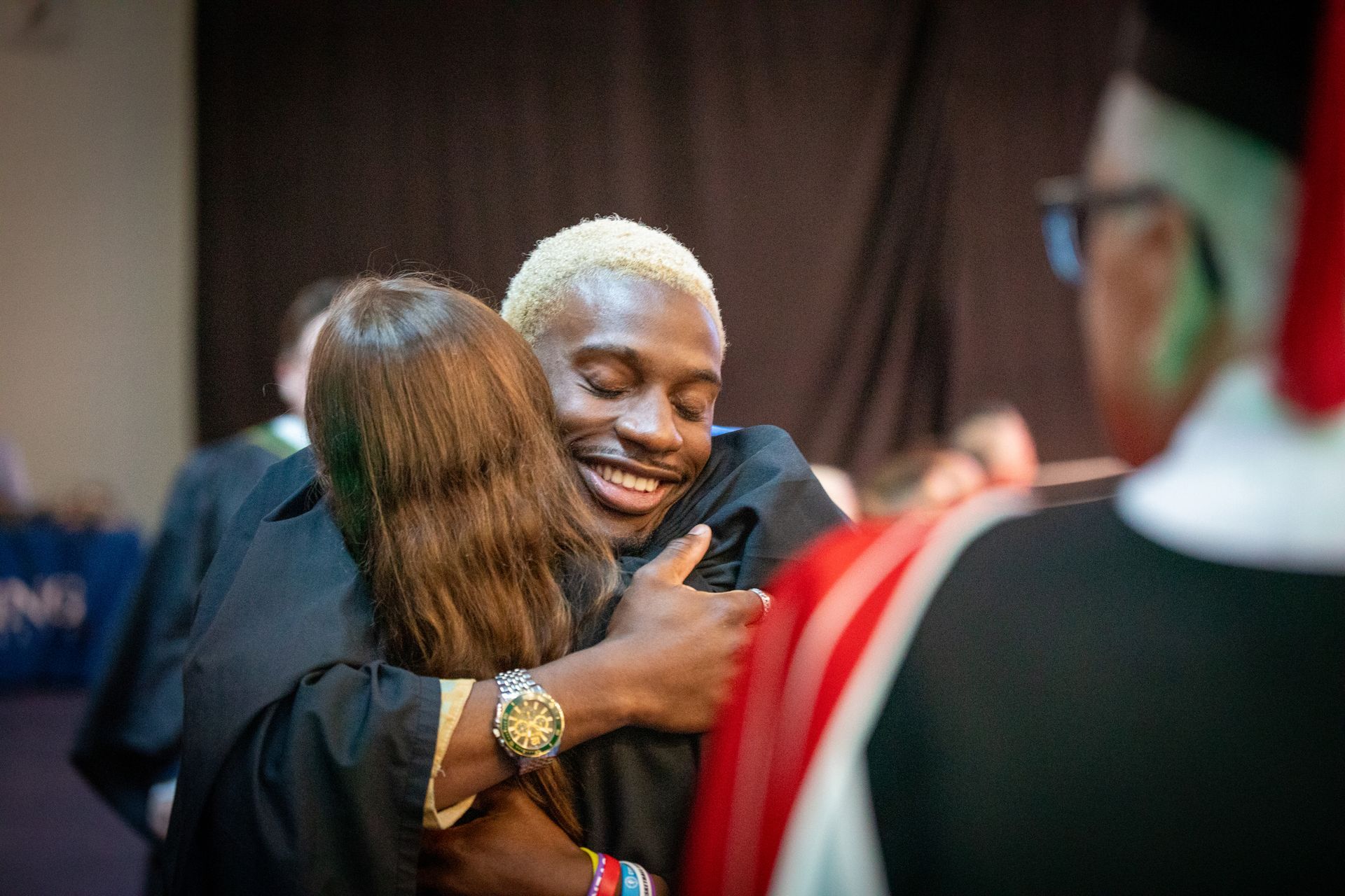 Two people hugging at an indoor event, smiling with blurred attendees in the background.