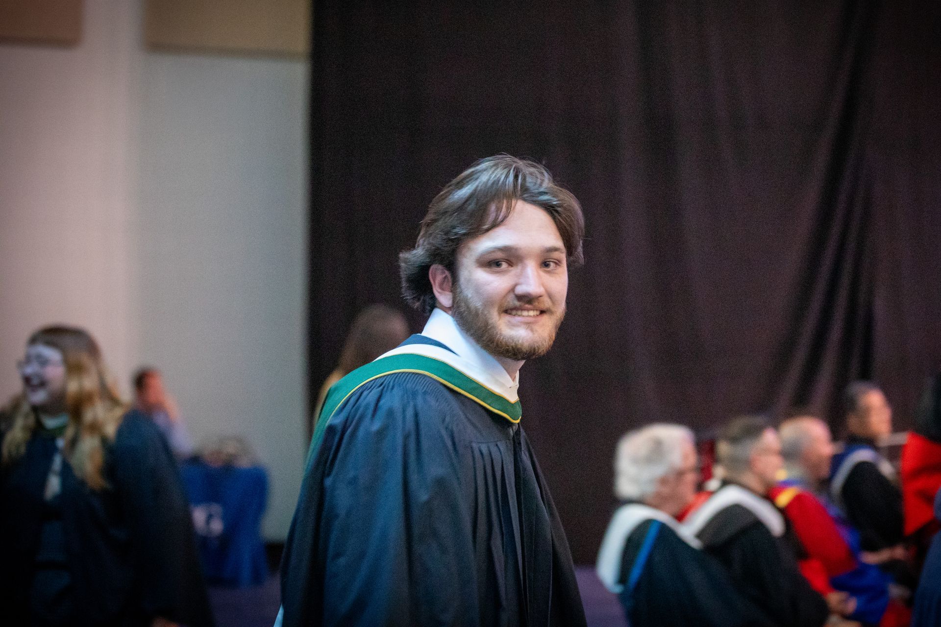 Man in black graduation gown with green trim smiling at ceremony, seated audience blurred behind him