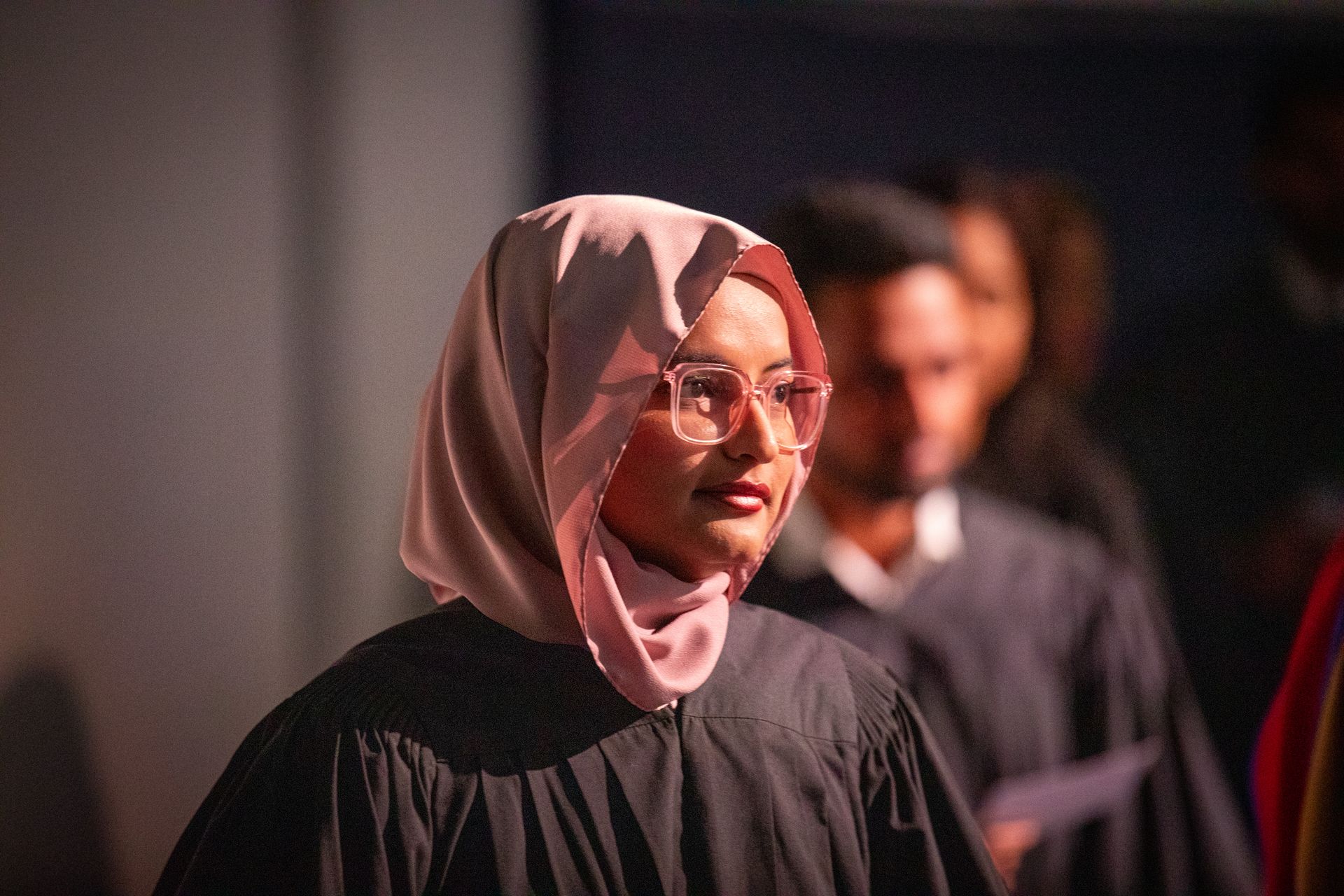 Woman in pink hijab and glasses standing in a dimly lit crowd