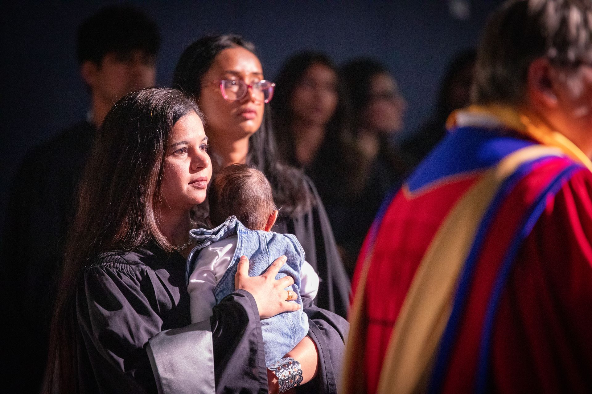 People standing in a dimly lit crowd, one holding a baby while facing a brightly robed figure.