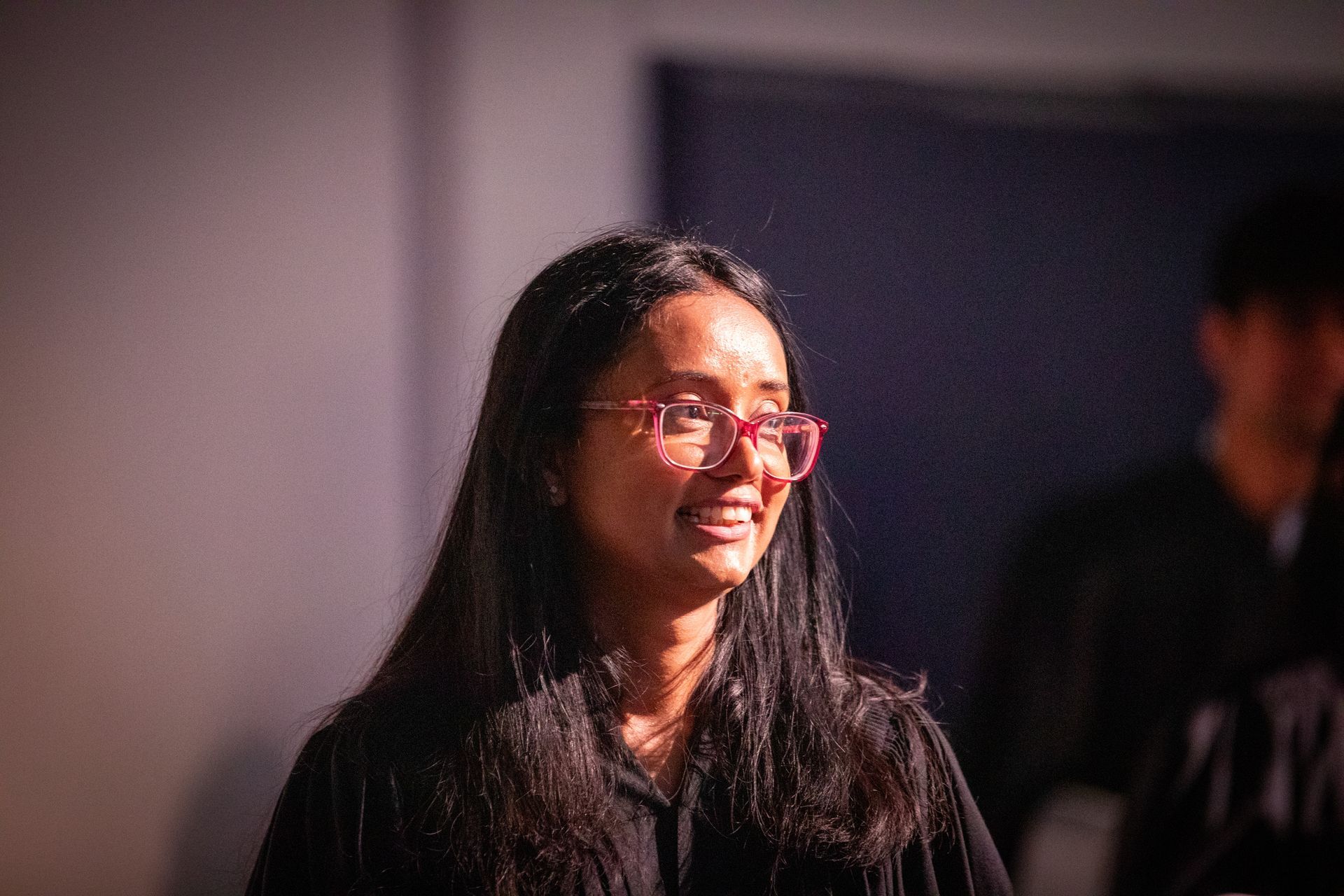 Woman with long dark hair and pink glasses smiling in a dimly lit indoor setting