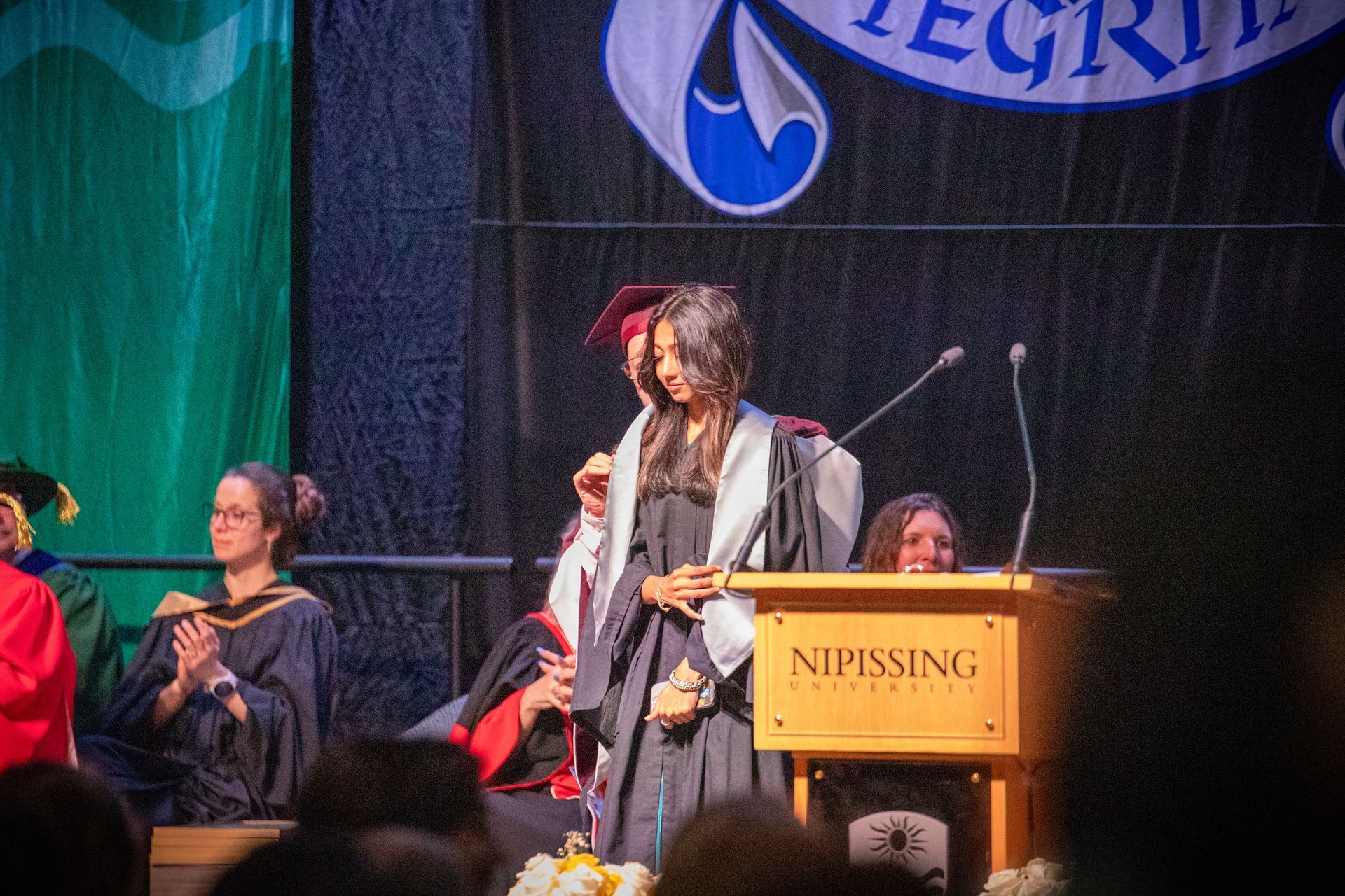 Speaker at podium on stage during ceremony, with seated attendees and a NIPISSING sign.