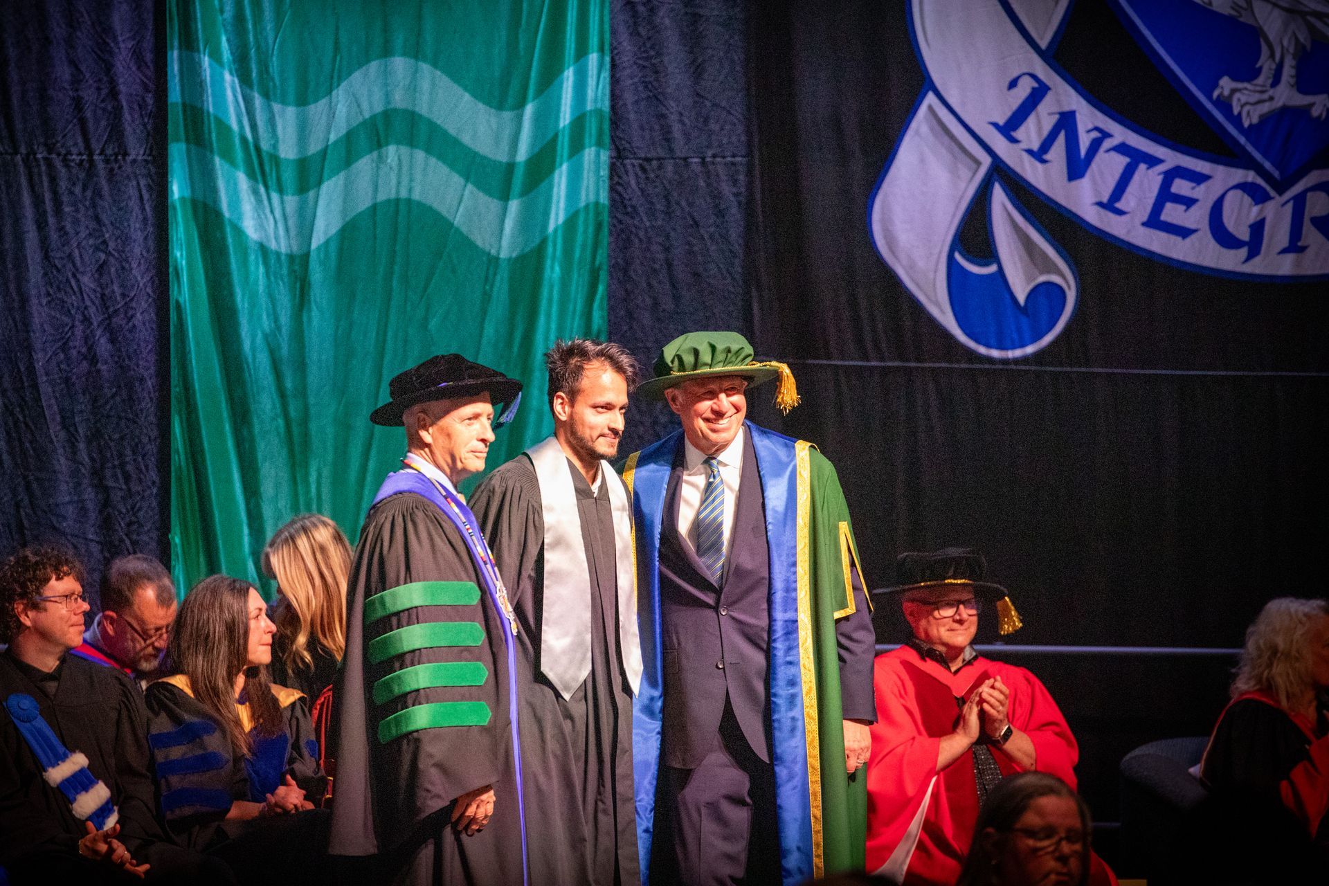 People in academic regalia onstage at a graduation ceremony, with seated audience in the background.