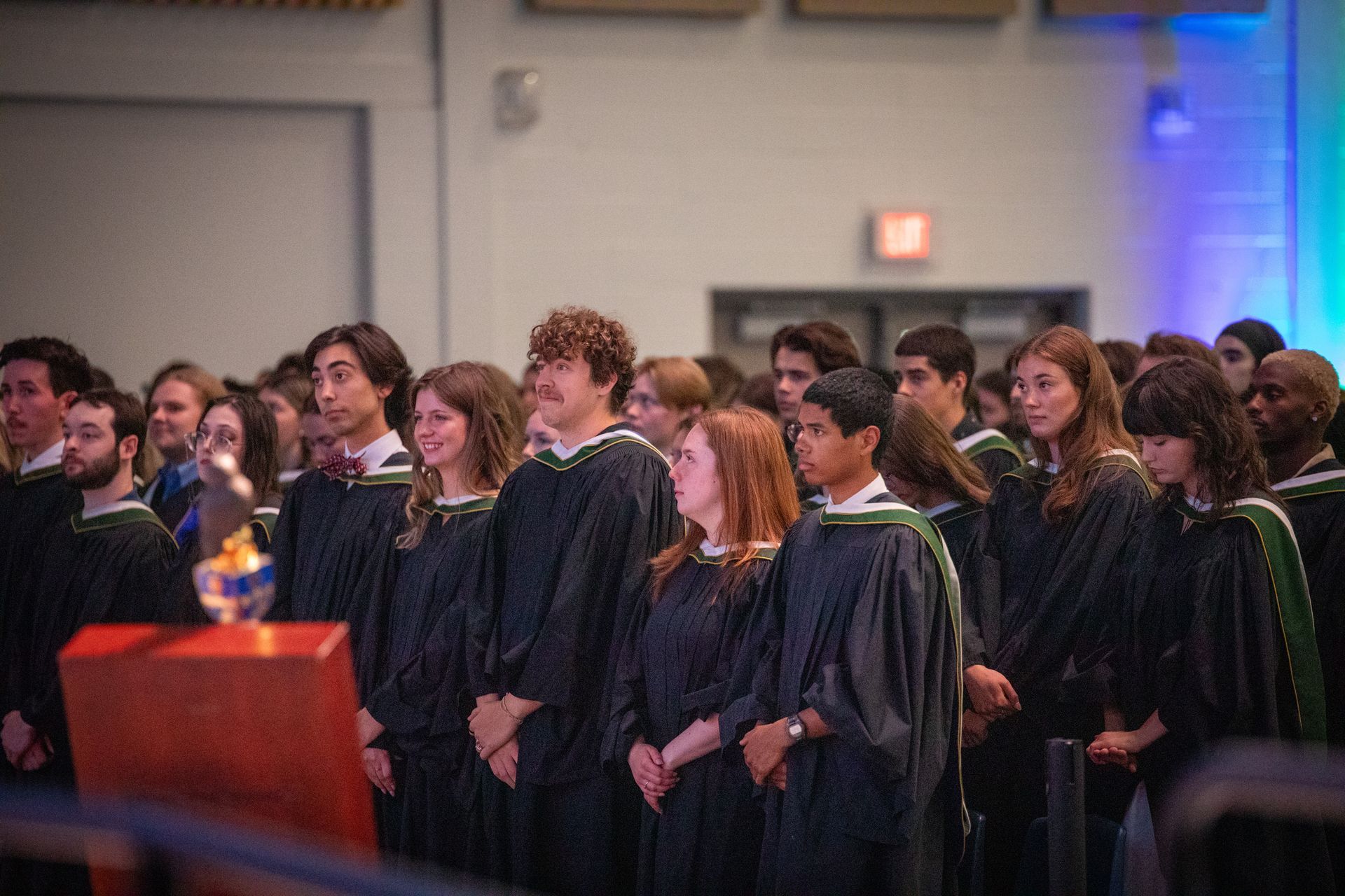 Graduates in black gowns standing in a ceremony hall, facing forward near a podium