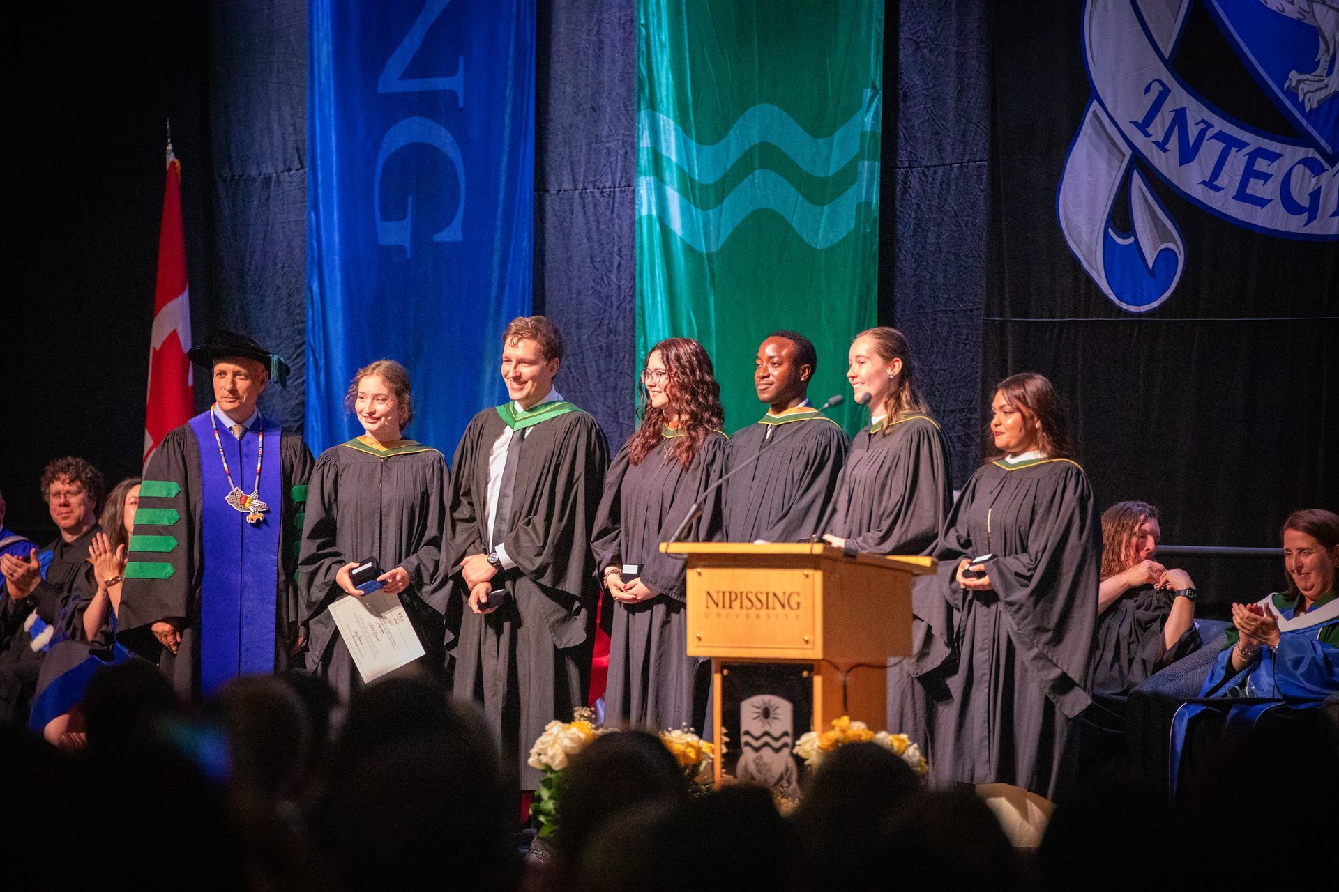 Graduates in black gowns stand onstage beside a podium at a commencement ceremony