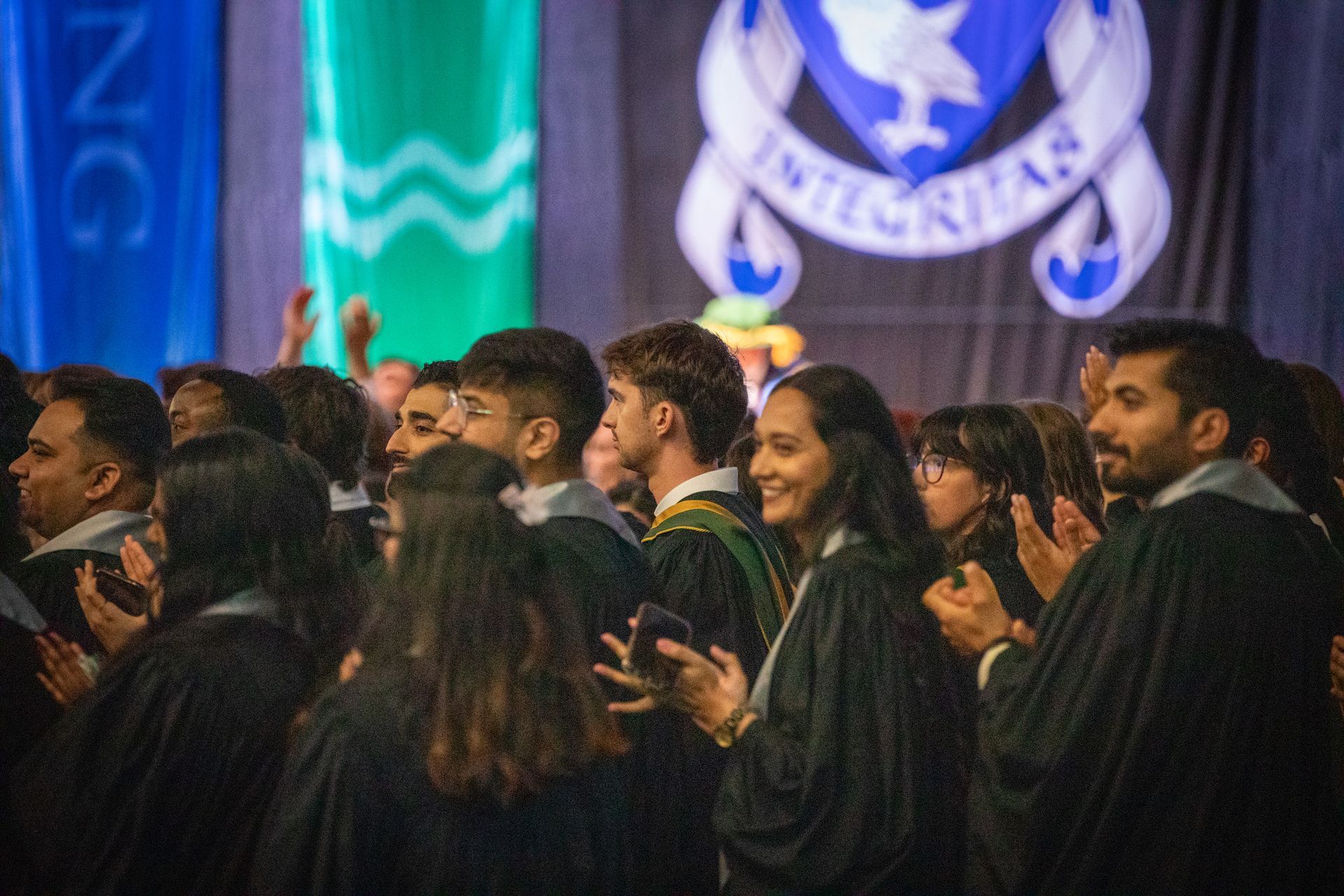 Graduates in black gowns stand together at a ceremony, smiling against blue and green banners.