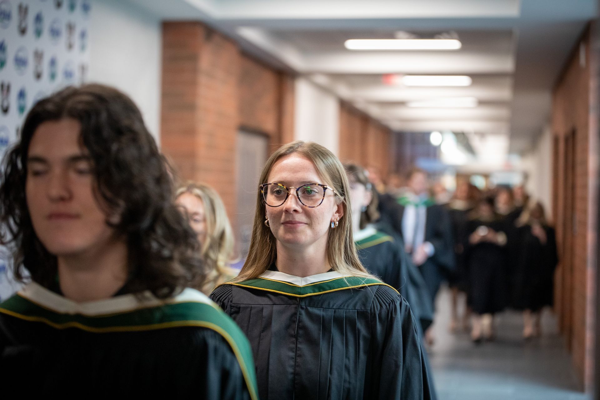 Graduates in black gowns walk down a school hallway, one smiling toward the camera.