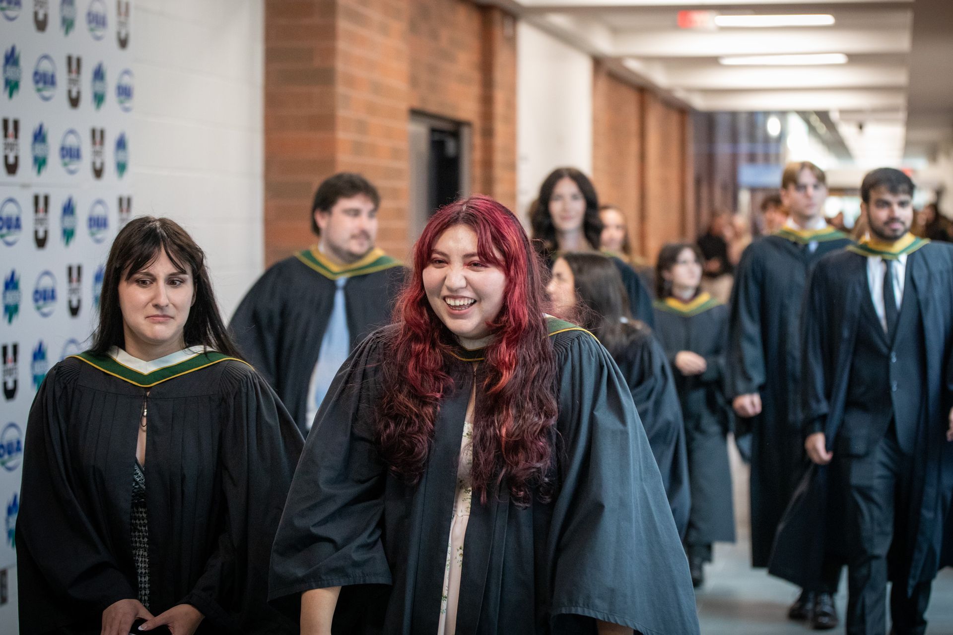Graduates in black gowns walking down a hallway, with one smiling red-haired graduate in front
