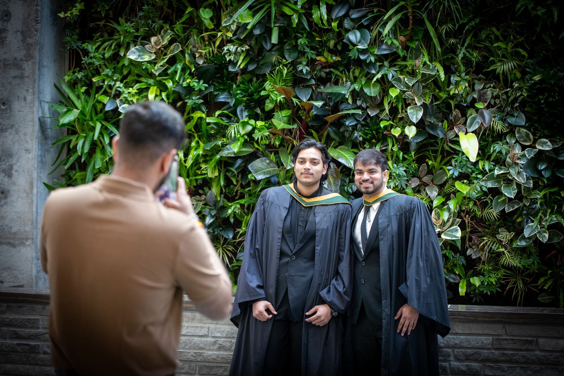 Two graduates in black gowns pose before a green wall while a photographer frames the shot.