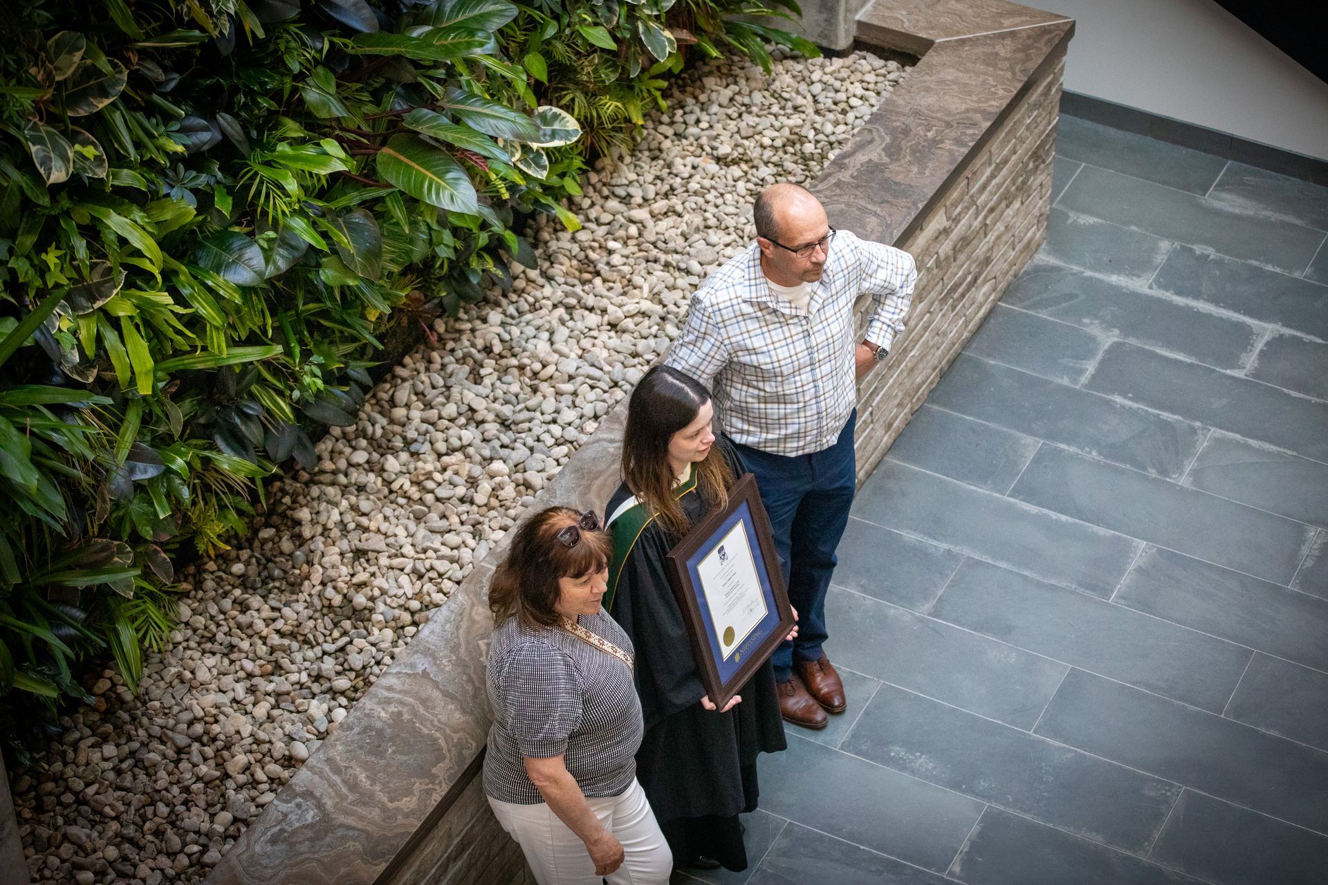 Three people standing on a stone walkway, one holding a framed certificate.