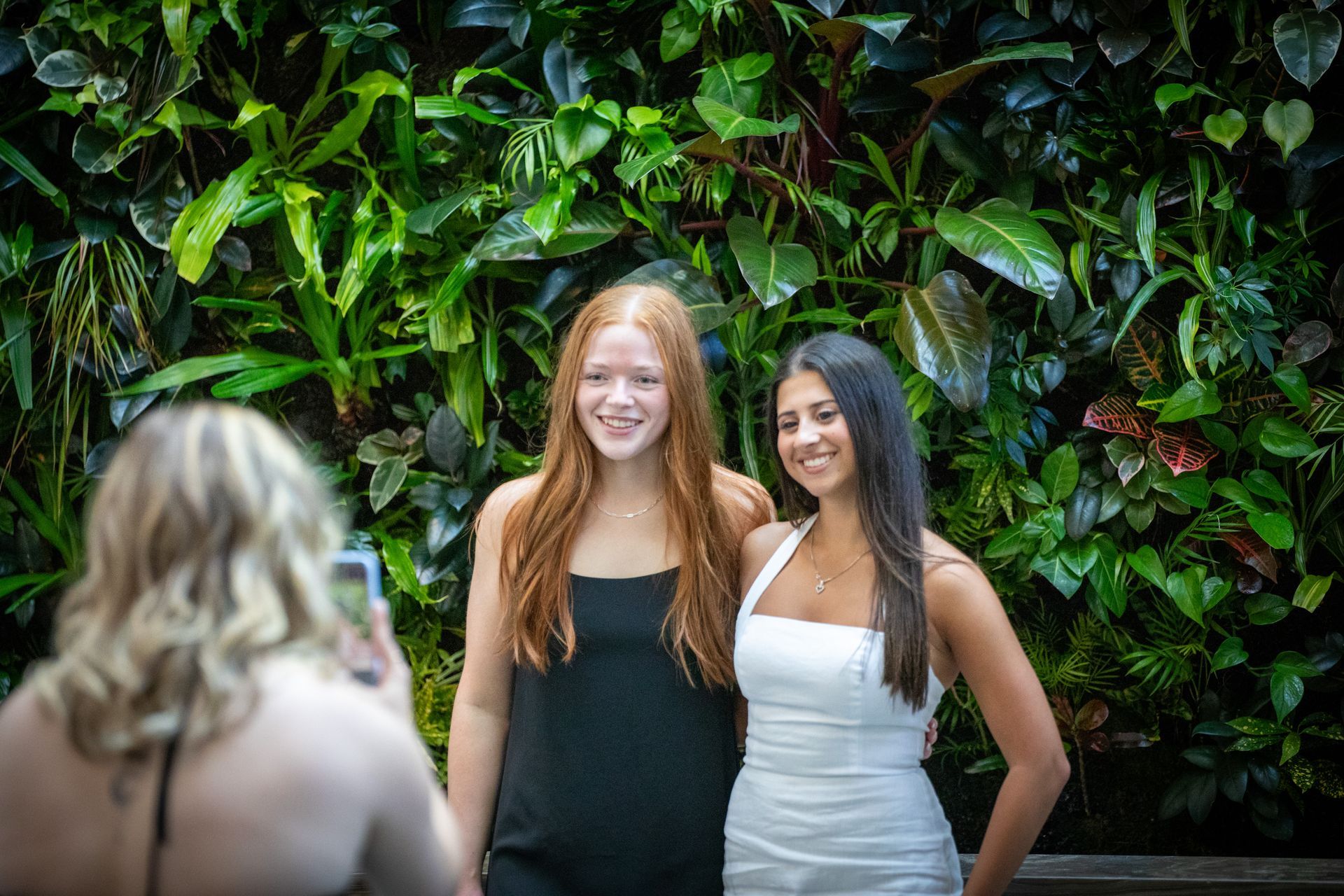 Two smiling women posing in front of a leafy green wall, with a blurred photographer in the foreground.