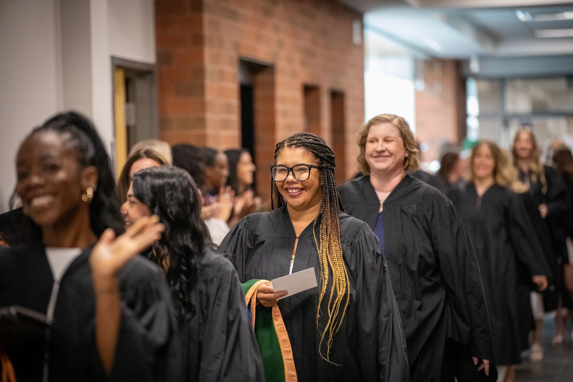 People in black graduation gowns walking in a hallway, smiling and carrying programs