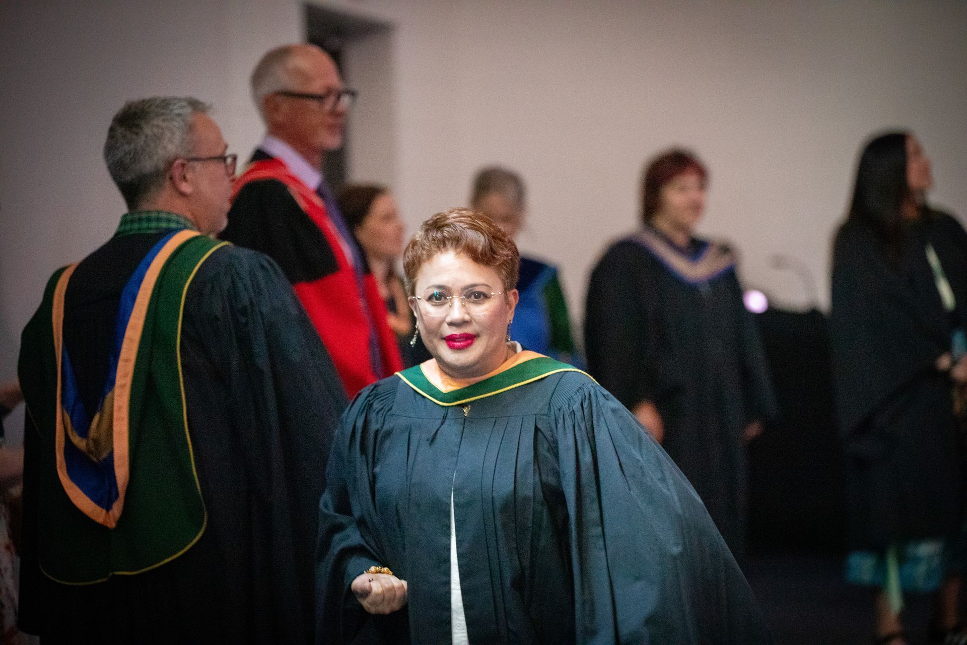 People in graduation robes standing indoors, with a woman in a green-trimmed gown in the foreground.