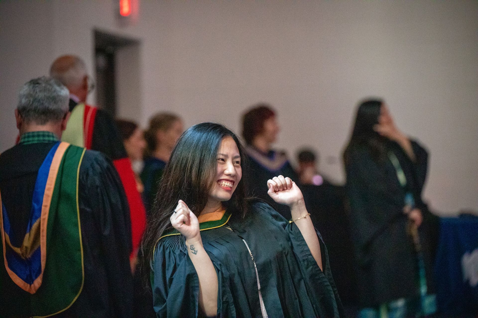 People in graduation gowns celebrating indoors, with one smiling woman raising her fists in the foreground