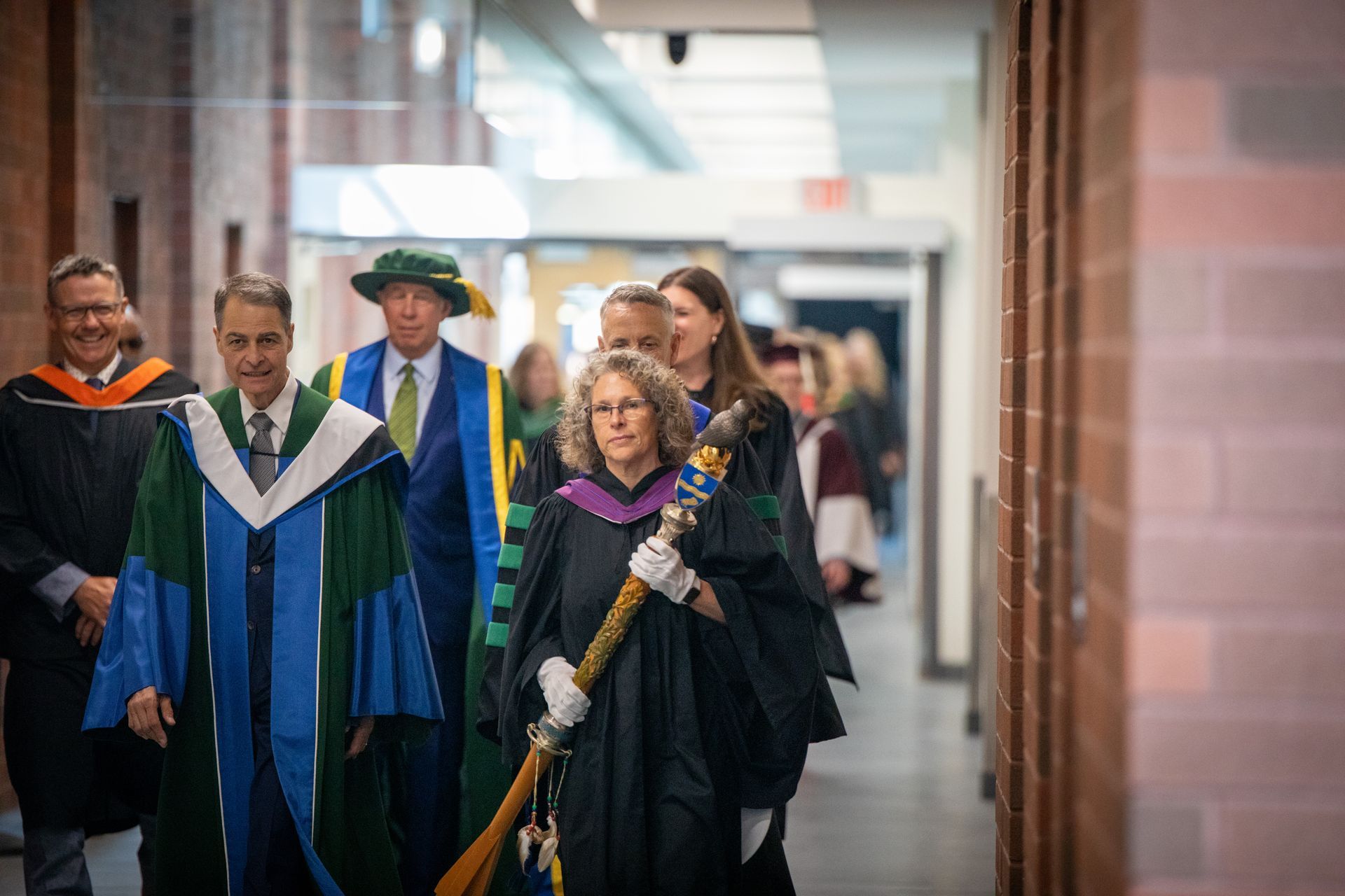 Officials in academic robes process through a hallway, one carrying a ceremonial staff.