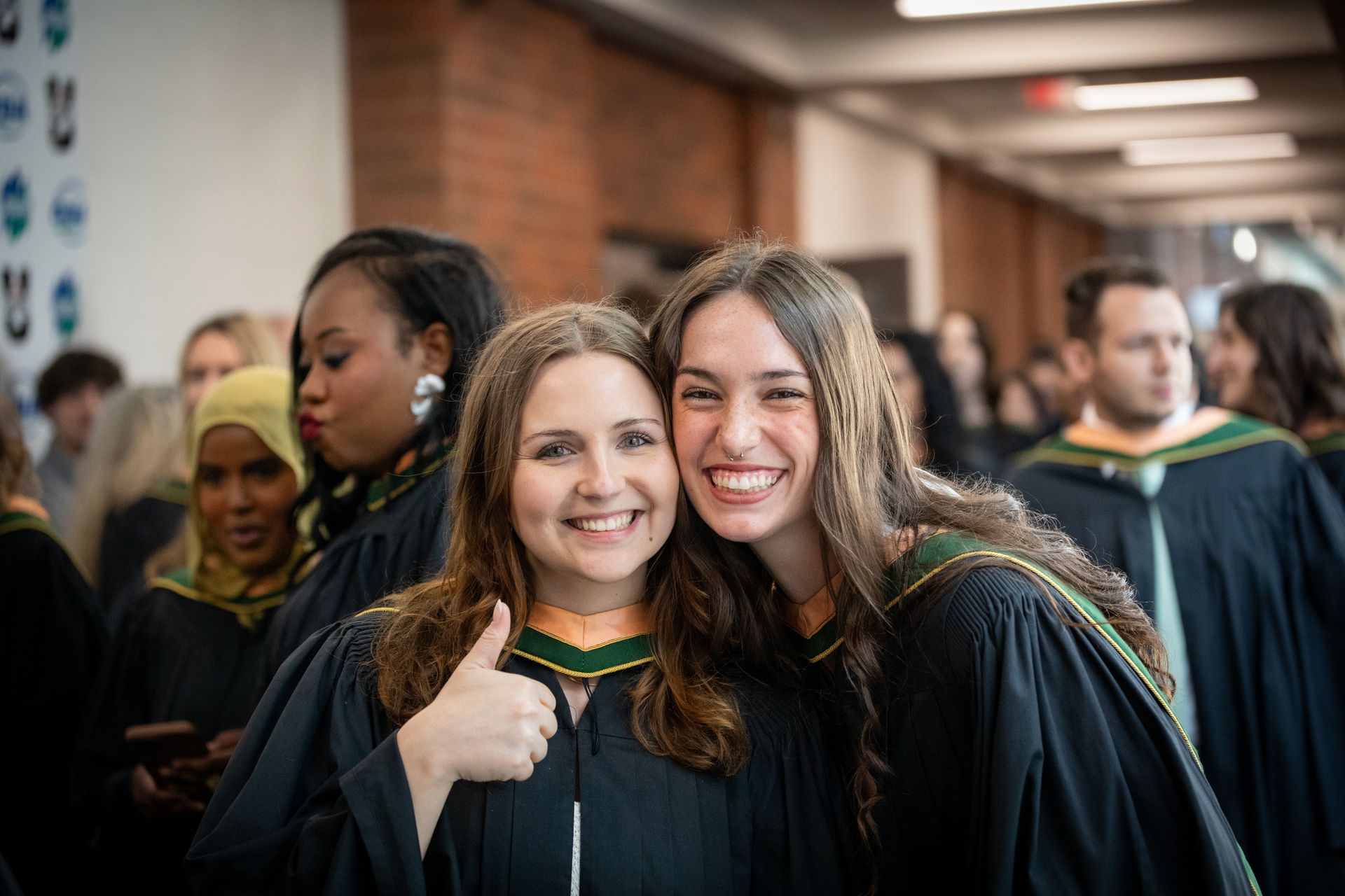 Two smiling graduates in black gowns pose together in a hallway, one giving a thumbs-up.