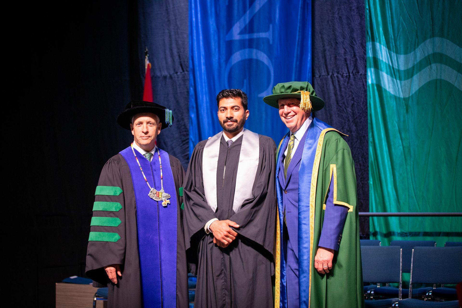Three people in caps and gowns posing on stage with diplomas and medallions