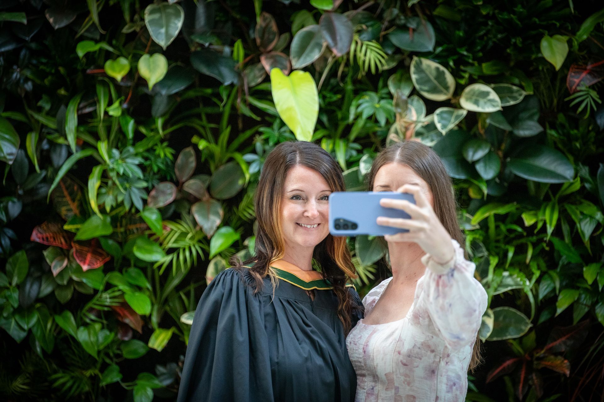 Two women taking a selfie in front of a lush green wall, one holding a blue phone and smiling