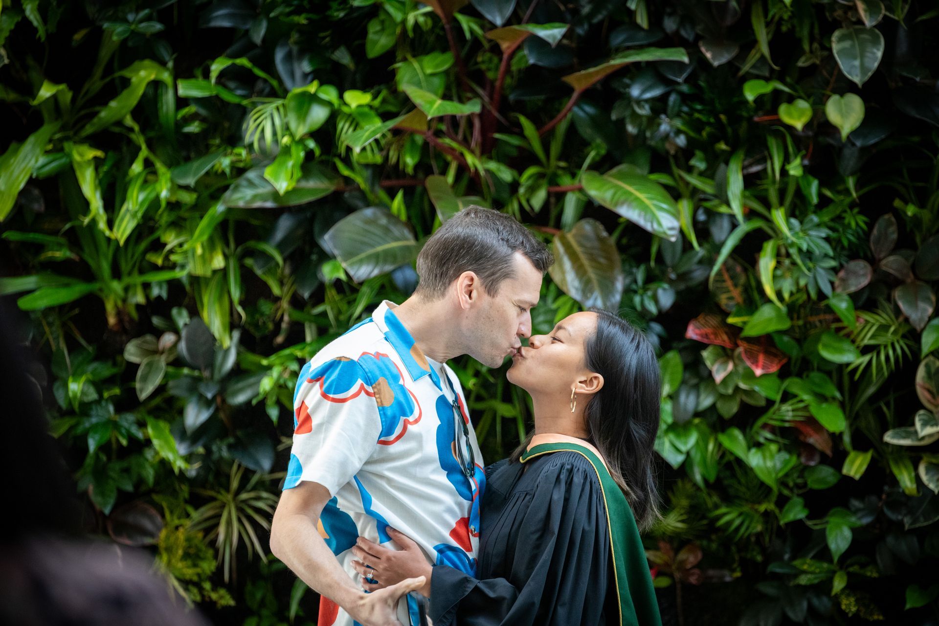 Couple kissing in front of a lush green wall of plants