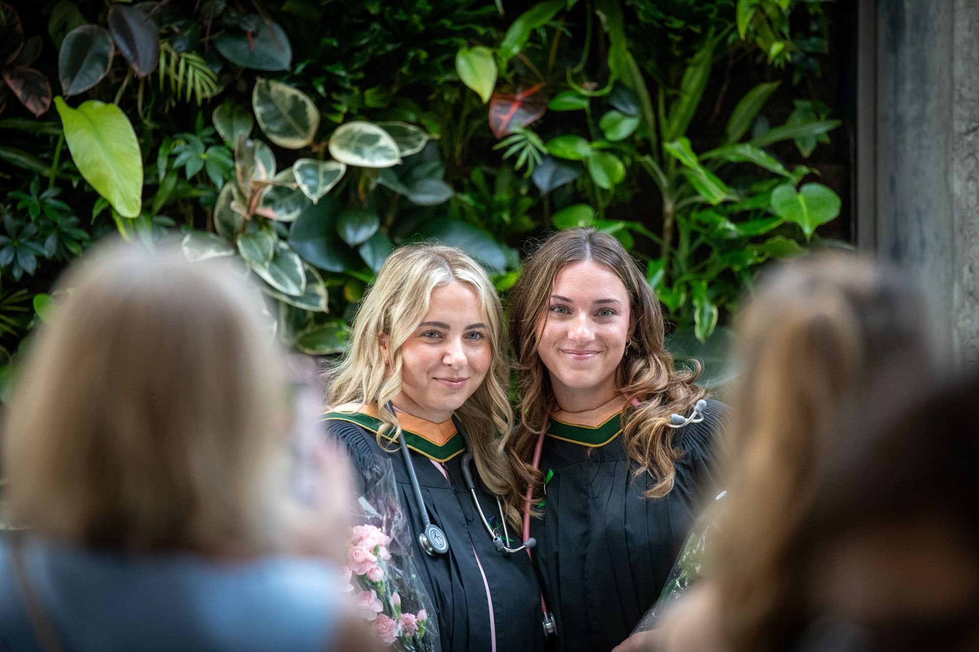 Two smiling women posing before a lush green wall, seen over blurred audience heads
